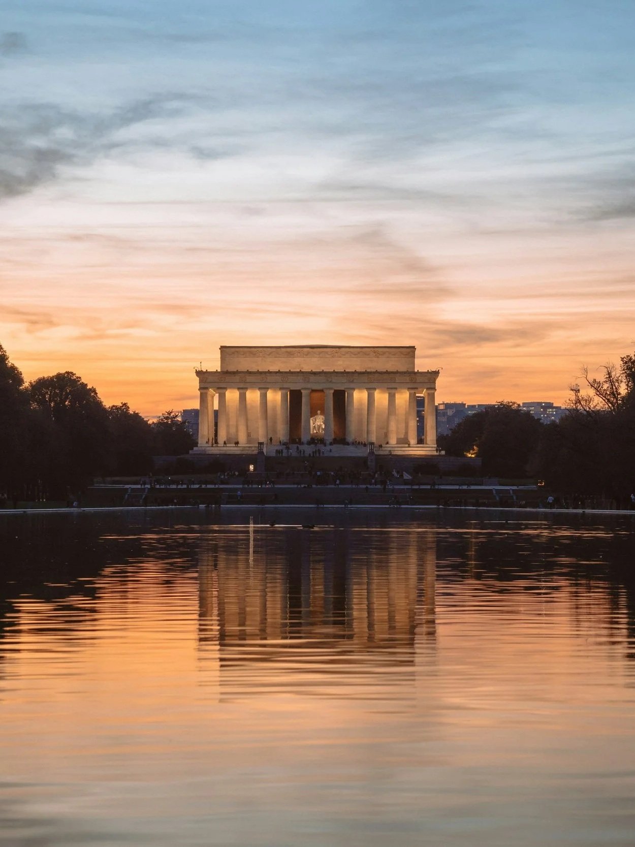 The Lincoln Memorial, a neoclassical monument, is pictured at sunset with an orange and blue sky, and its reflection is visible in the water in the foreground.