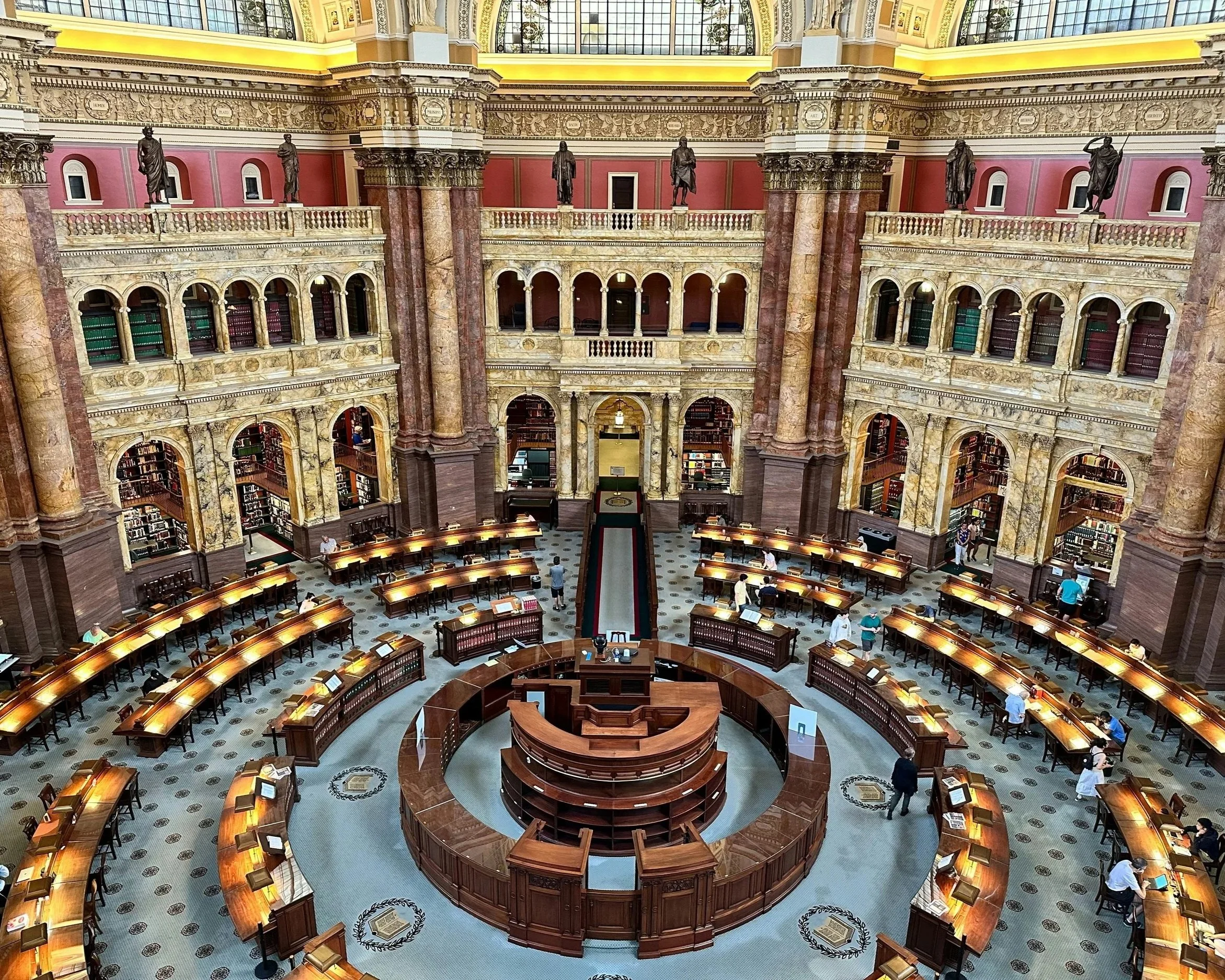 Interior of a grand historic library or archive with multiple levels of bookshelves, ornate architecture, mounted statues, and visitors seated at tables.