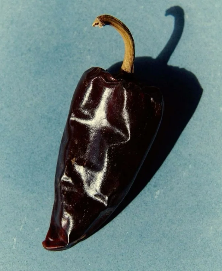 A dark chili pepper with a face-like texture, casting a shadow on a blue surface.
