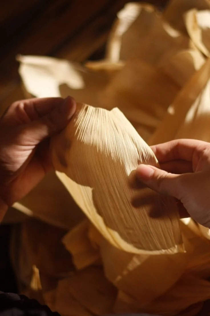 Hands peeling back a banana leaf, revealing its textured surface, with more banana leaves visible in the background.
