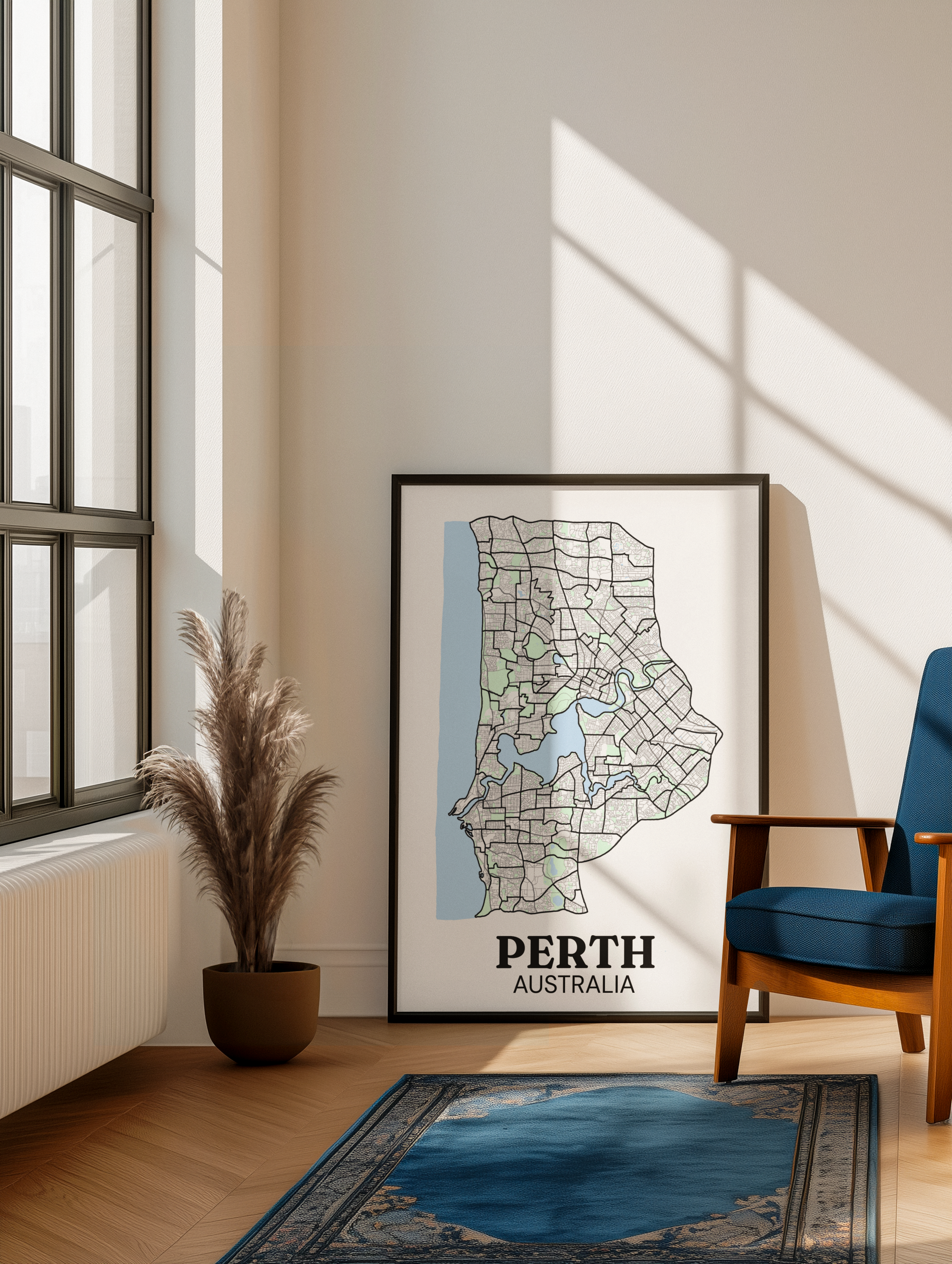 Black-framed colour street map print of Perth, Australia leaning against a wall in a sunlit living room with a pampas grass plant and blue Persian rug.