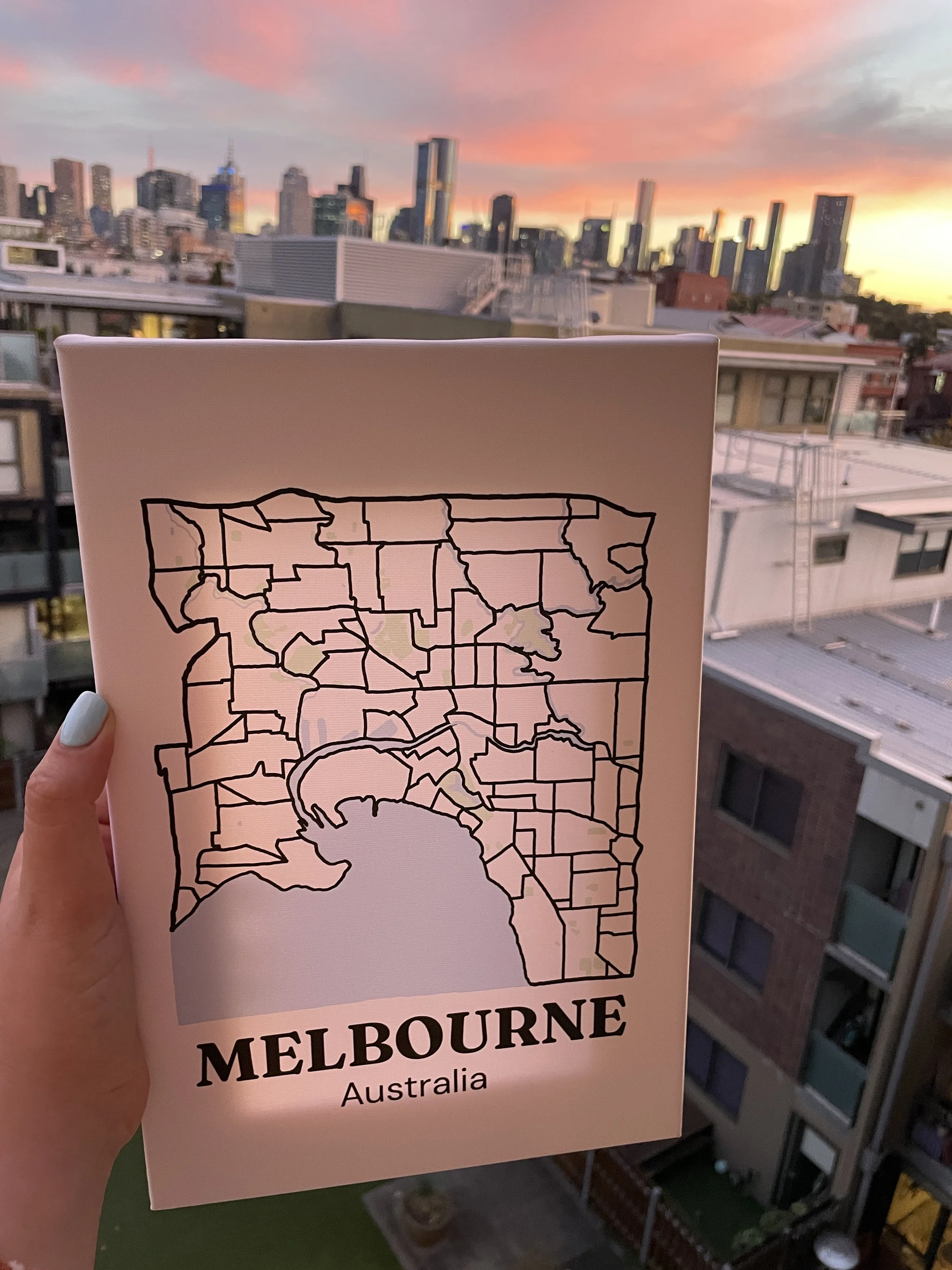 Blank hand-drawn Melbourne suburb map canvas held up against a sunset city skyline, showing the outline design ready to be painted in as suburbs are visited