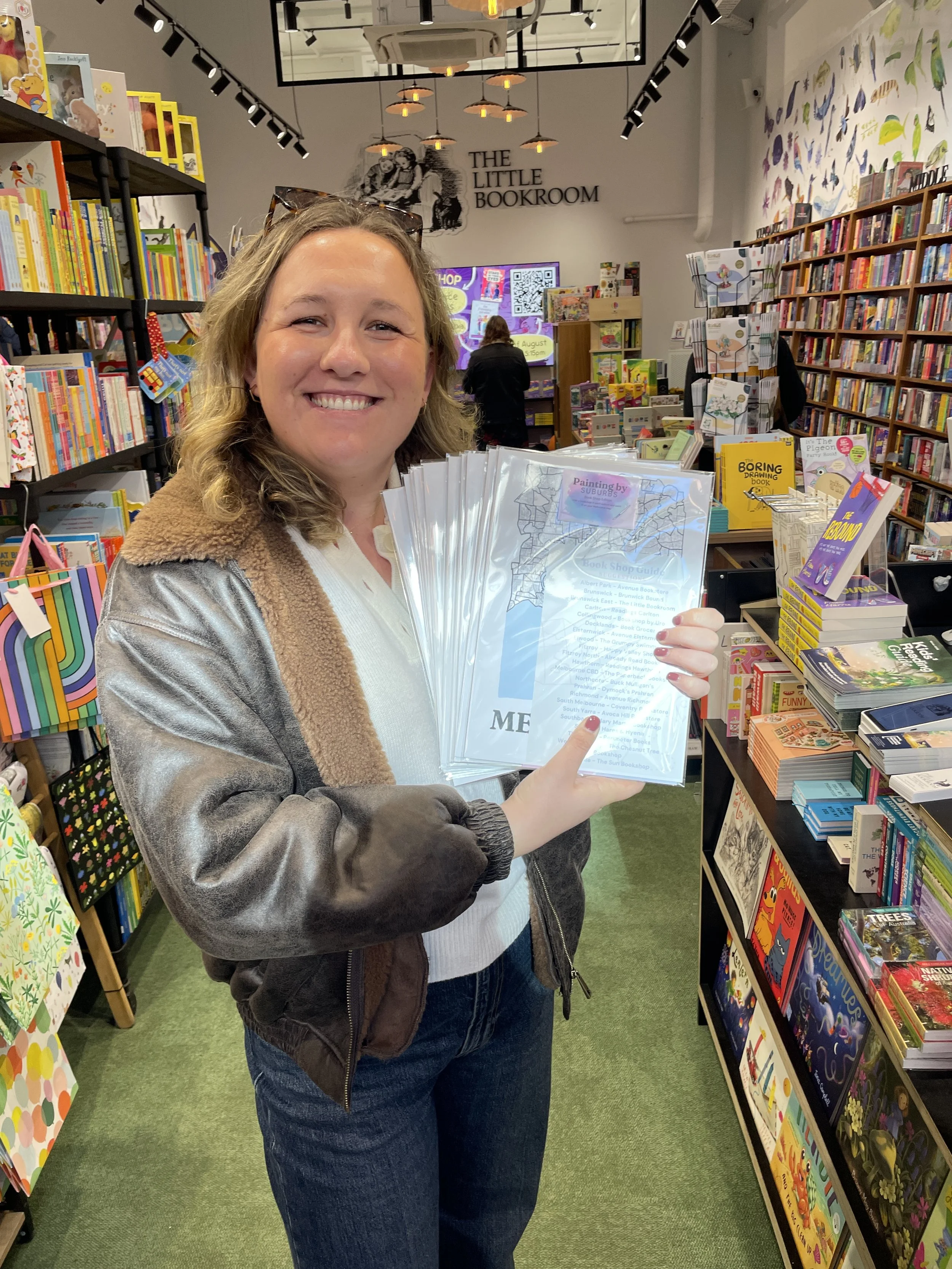 Annie smiling and holding a stack of Painting by Suburbs Book Shop Guides inside The Little Bookroom, a children's bookshop with colourful shelves.