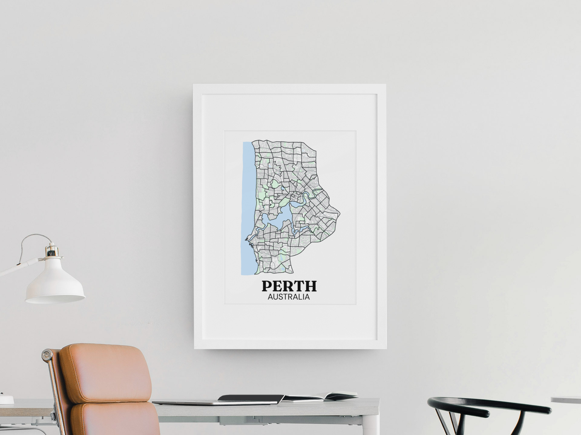 White-framed colour street map print of Perth, Australia hung on a wall above a desk with a tan leather chair.