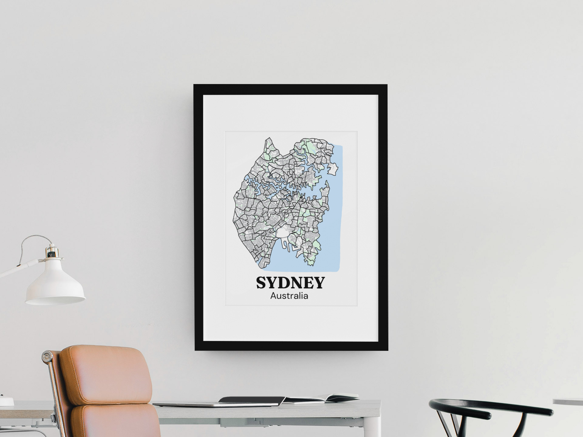 Framed map of Sydney, Australia, hanging on a white wall in an office setting with a desk, a tan chair, and a white desk lamp visible.