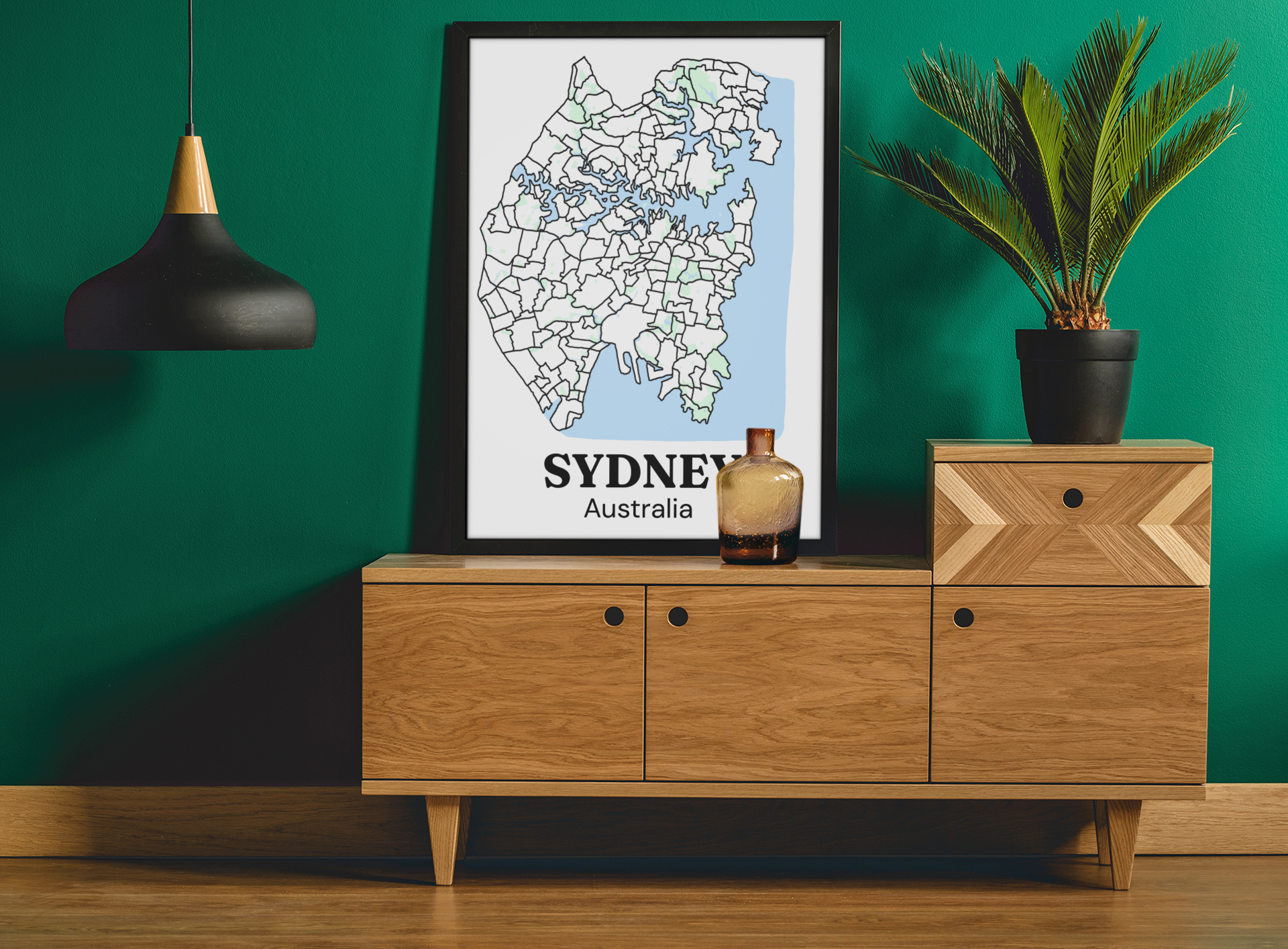Black-framed bare Sydney suburb map print leaning on a wooden sideboard against a deep green wall with a potted palm.