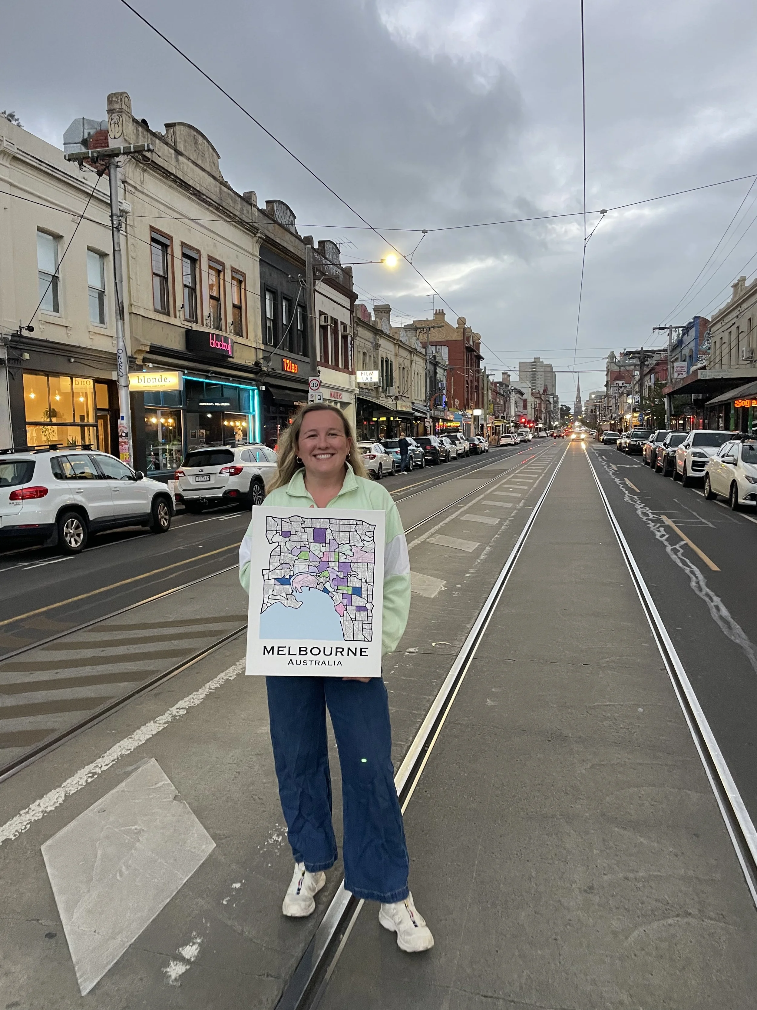 A woman standing on a city street tram track holding a sign with a map of Melbourne, Australia, smiling at the camera. The street is lined with parked cars and historic buildings, with a cloudy sky overhead.