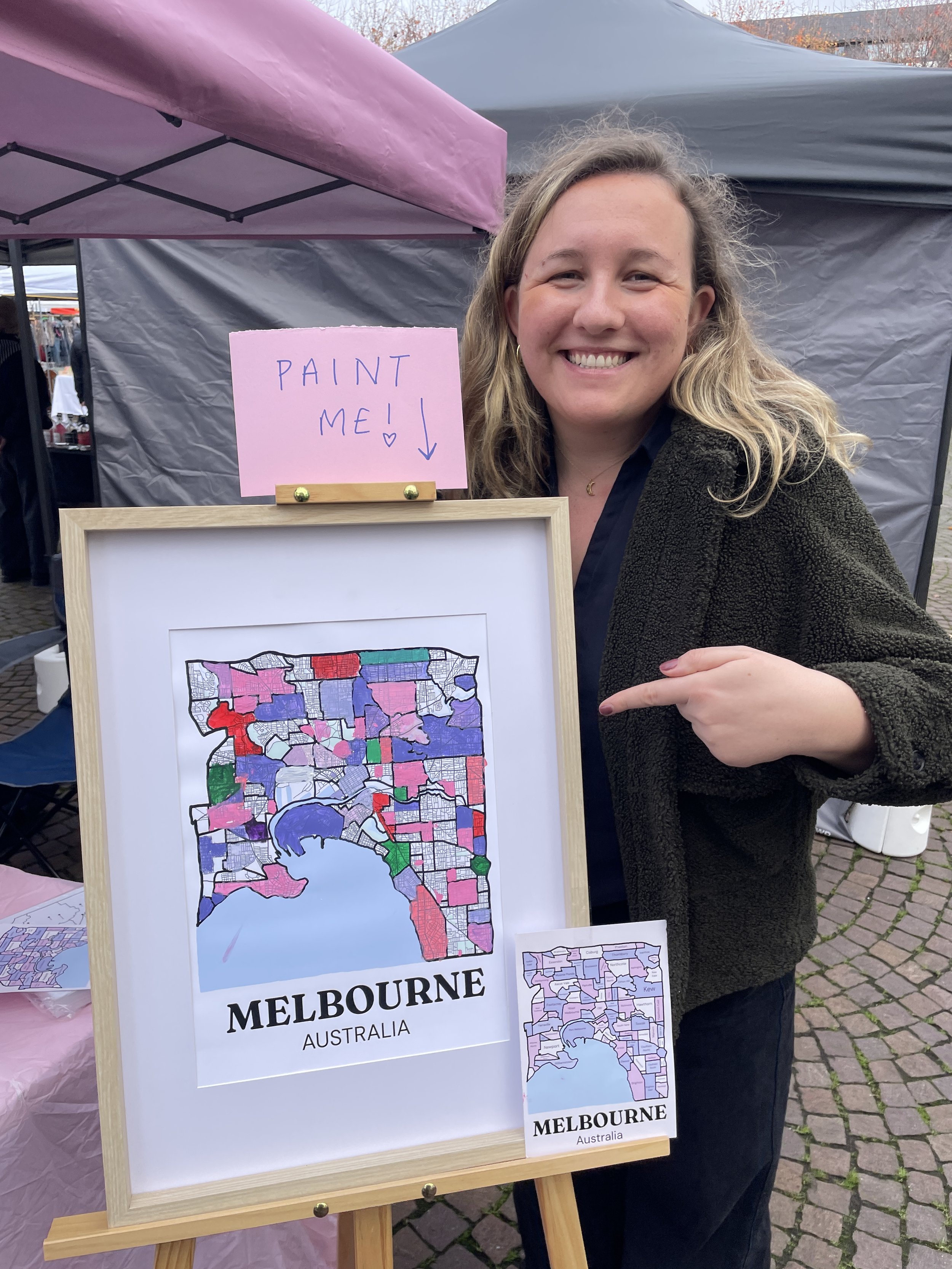 Painting by Suburbs founder smiling and pointing at a colourfully hand-painted Melbourne suburb map on an easel, with a 'Paint Me' sign, at an outdoor market stall.