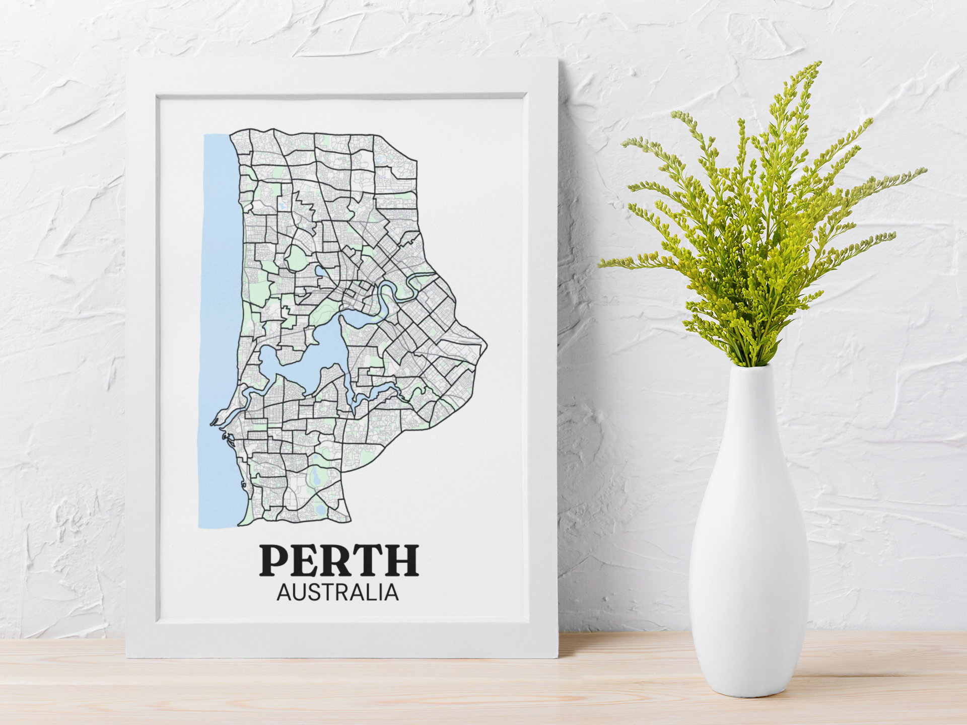 White-framed colour street map print of Perth, Australia leaning against a textured white wall beside a white vase with greenery.