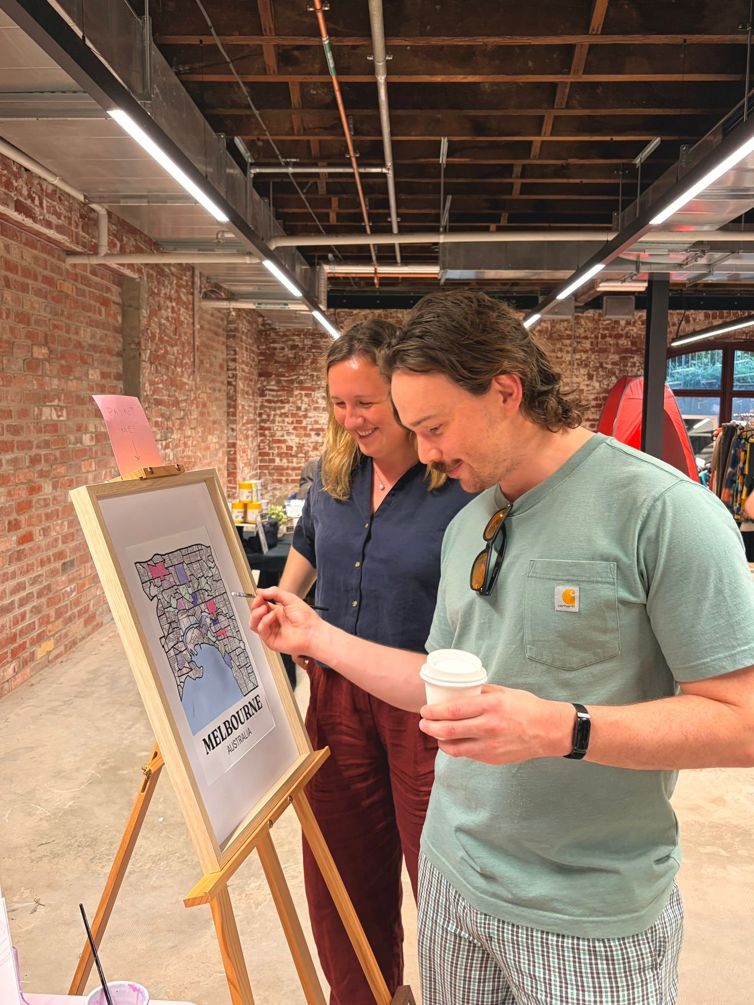 Market visitor painting in suburbs on a framed Melbourne suburb map displayed on an easel with a 'Paint Me' sign, demonstrating the interactive hand-painted map concept