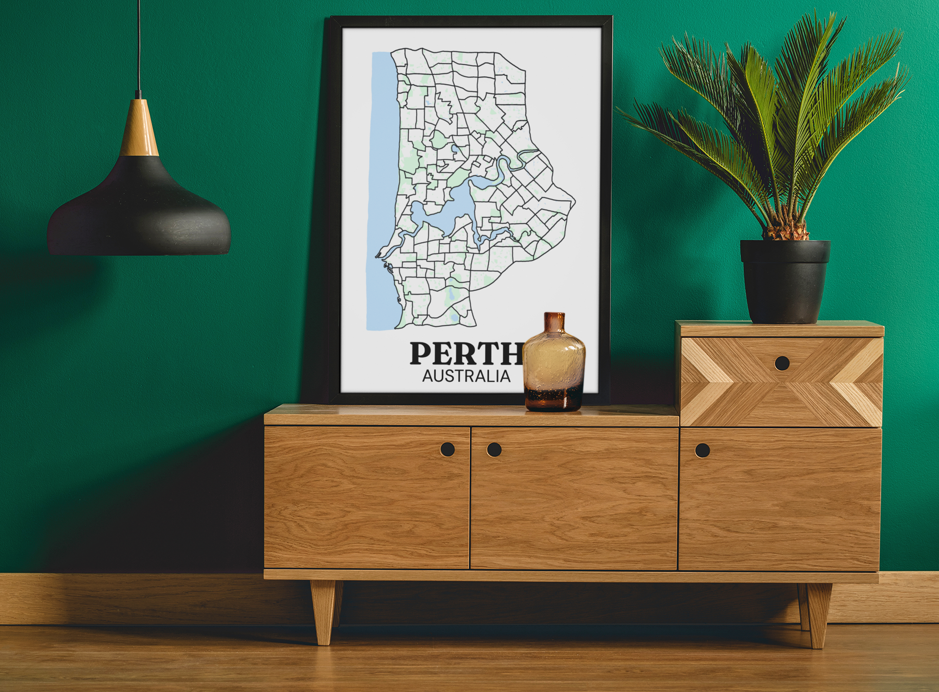 Black-framed suburb boundary map print of Perth, Australia displayed on a wooden sideboard against a green wall with a potted palm.