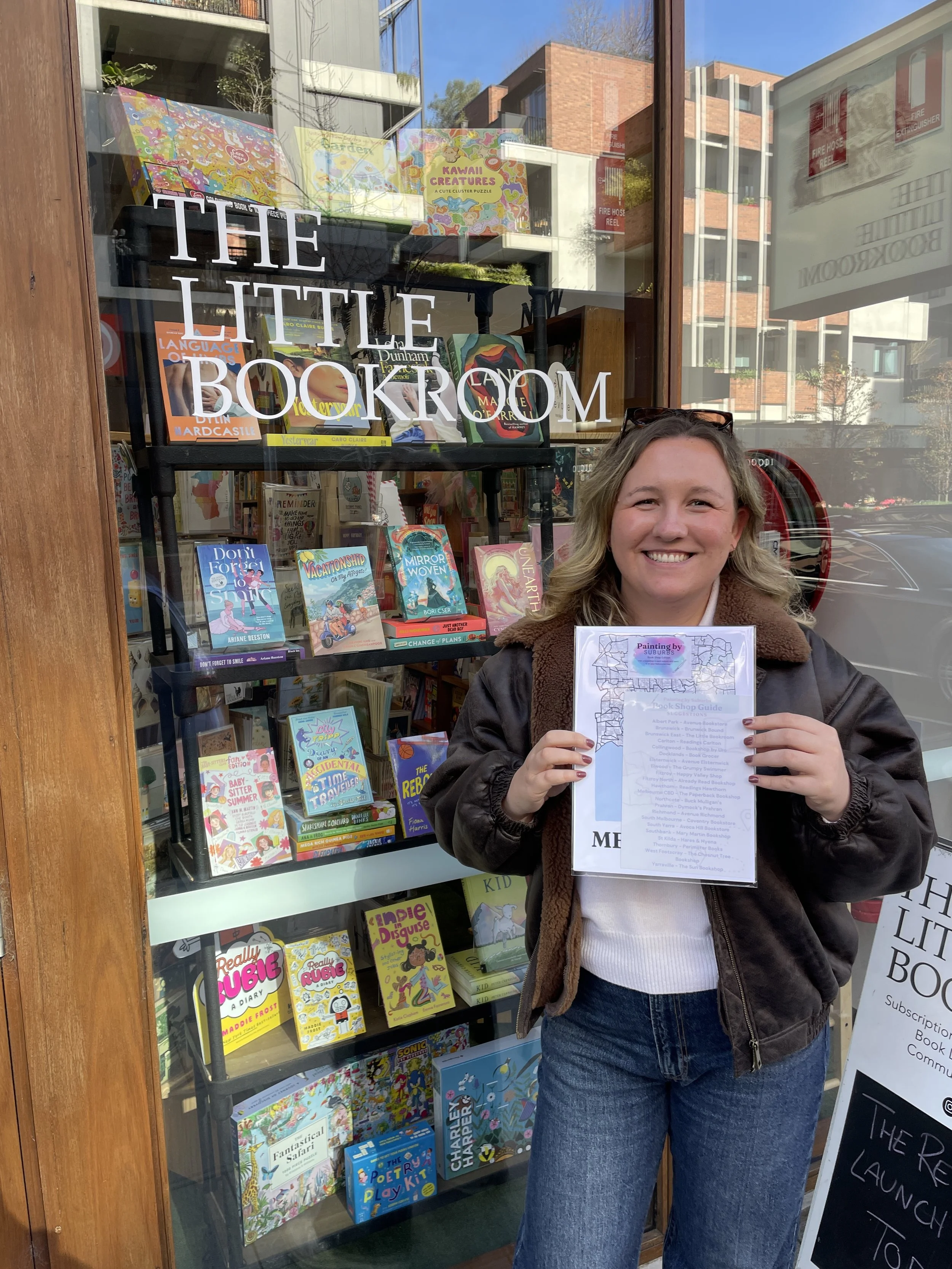 Annie founder smiling and holding the Painting by Suburbs Bookshop Guide outside The Little Bookroom, an independent bookshop with a window display of books and puzzles.