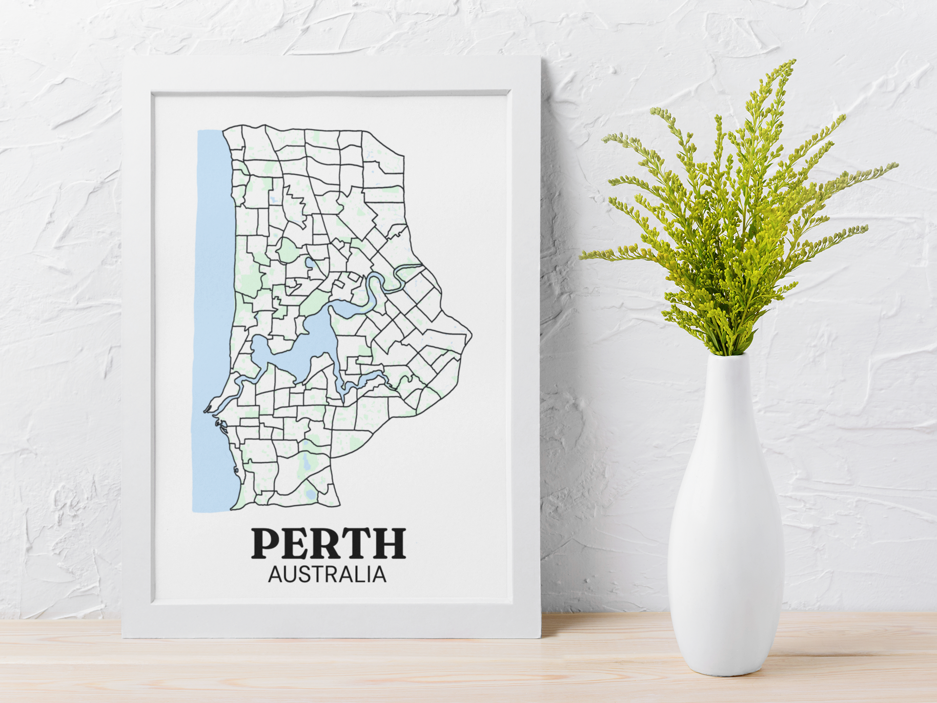 White-framed suburb boundary map print of Perth, Australia leaning against a wall beside a white vase with greenery.