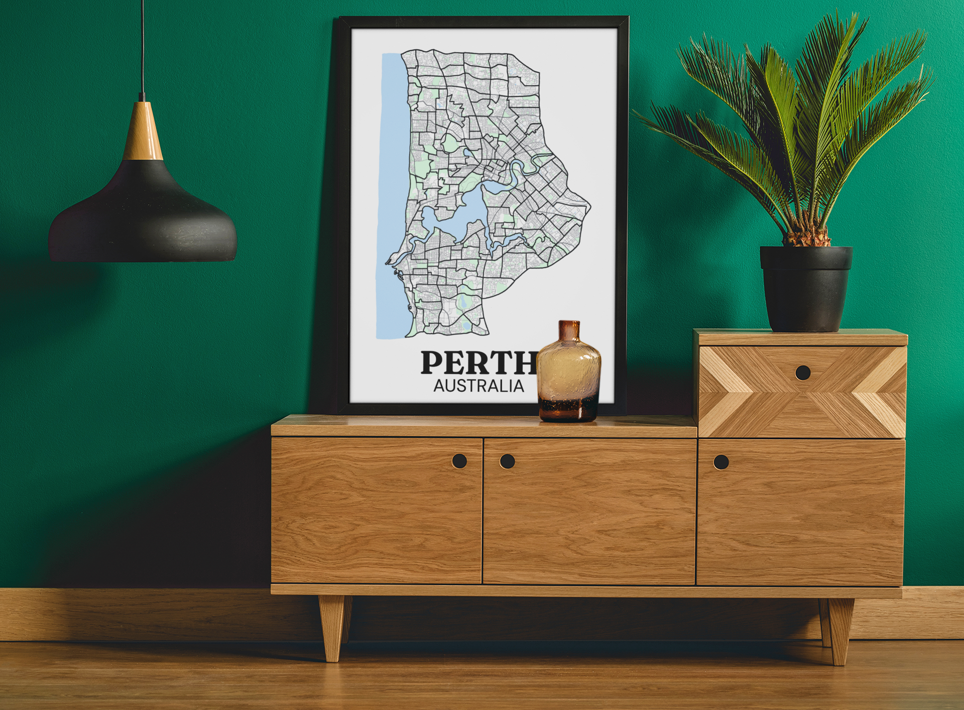 Black-framed colour street map print of Perth, Australia displayed on a wooden sideboard against a green wall with a potted palm.