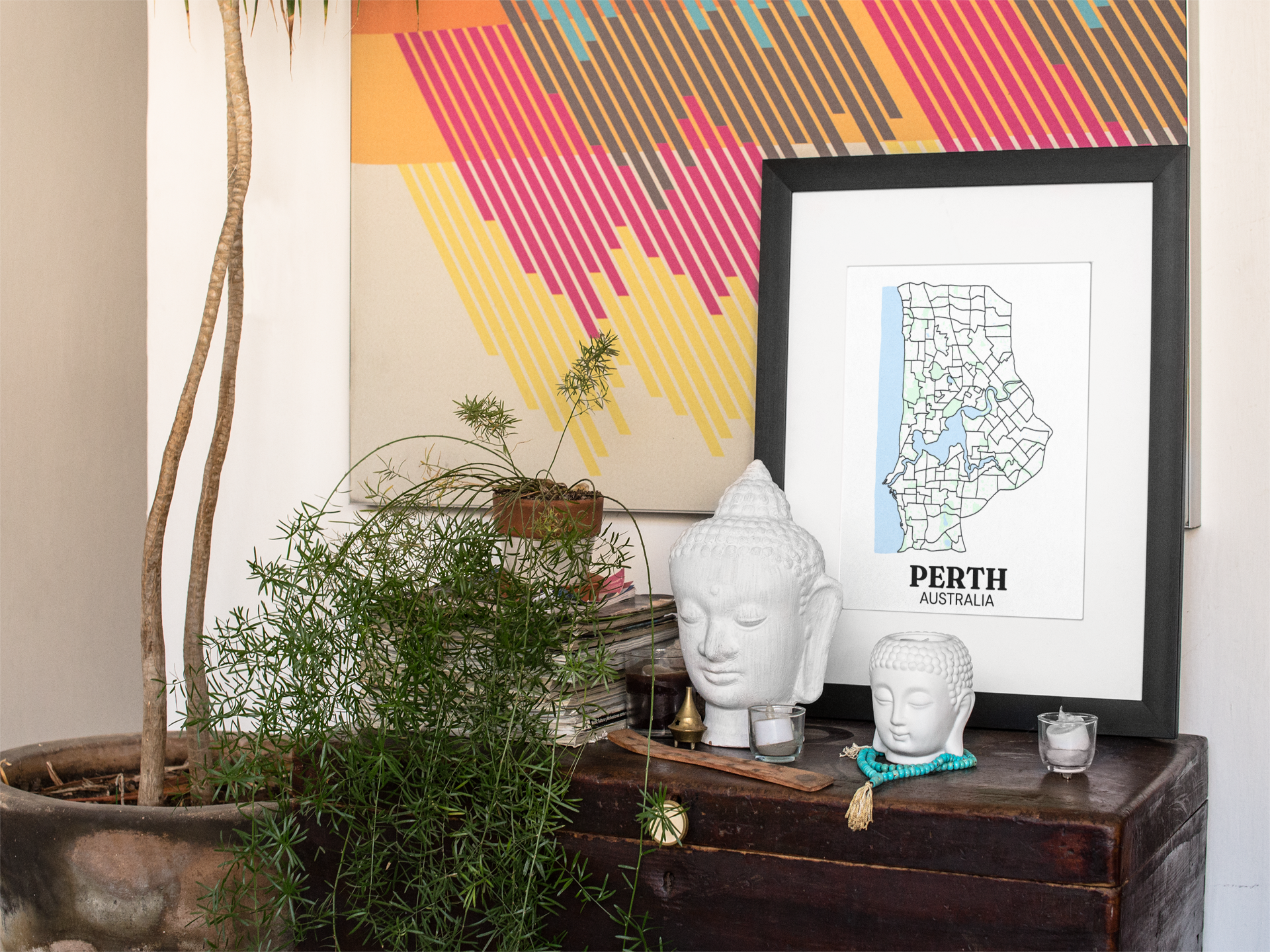 Black-framed suburb boundary map print of Perth, Australia displayed on a dark wooden chest beside ceramic Buddha head ornaments and potted plants.