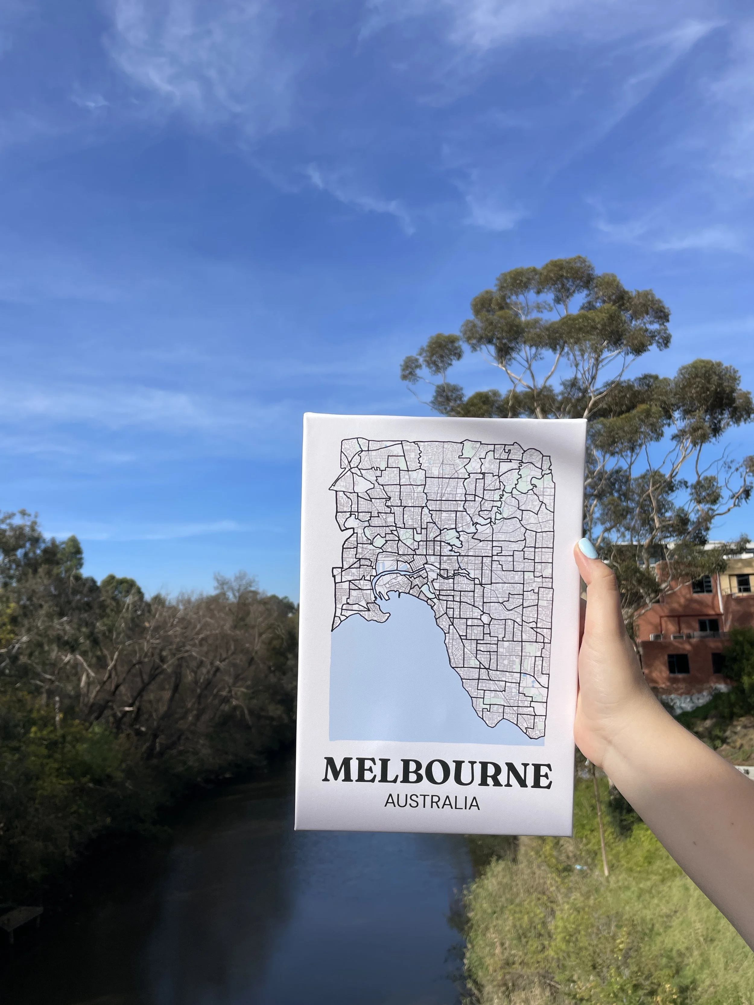 Hand-drawn Melbourne suburb map canvas held up against a blue sky and river, showing the full suburb outline design with parks and coastline detail