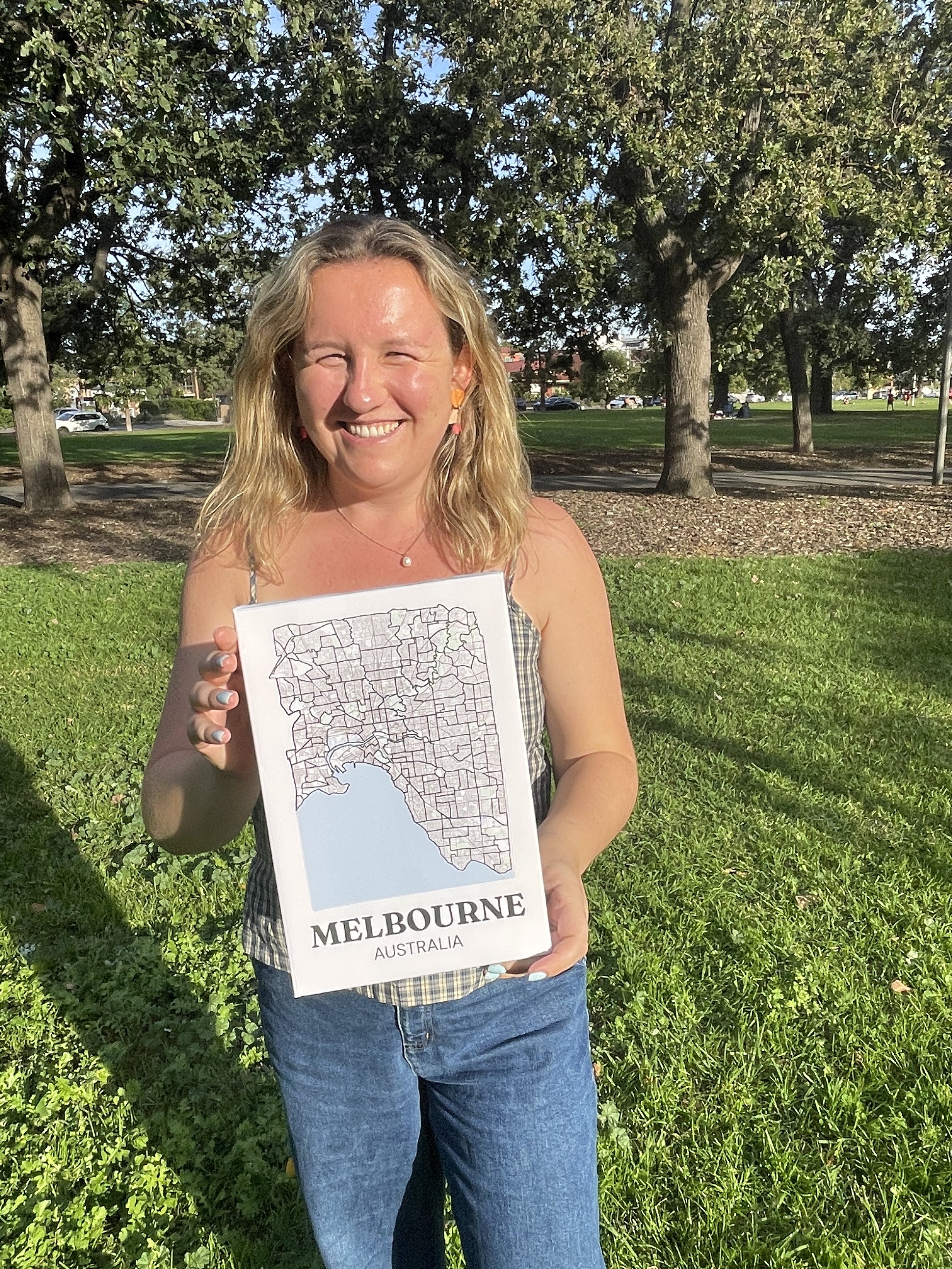 Painting by Suburbs founder holding up an unframed hand-drawn Melbourne suburb map print outdoors in a park, showing the full suburb outline design with coastline detail