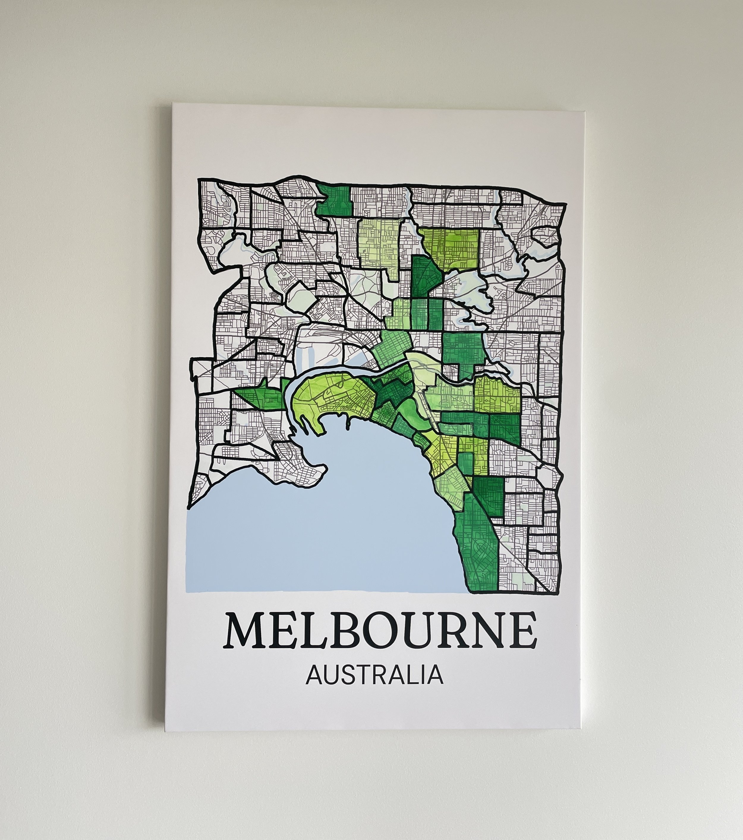 Canvas Melbourne suburb map hanging on a wall, with multiple suburbs hand-painted in shades of green marking places visited, bay coastline visible at the bottom.