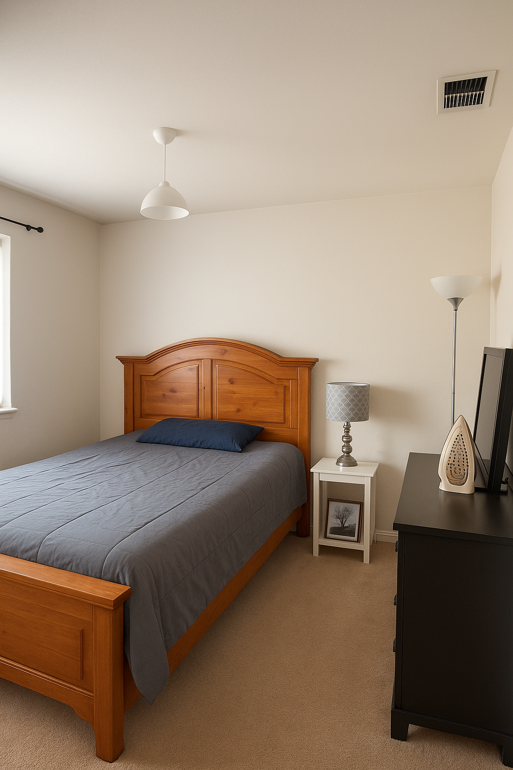 A bedroom with a wooden bed frame, a gray bedspread, and a blue pillow. There is a small white nightstand with framed photo, a gray table lamp, and a picture frame. A black dresser with a decorative iron and a TV is visible. A floor lamp, ceiling light, air vent, and a window with a curtain rod are also in the room.