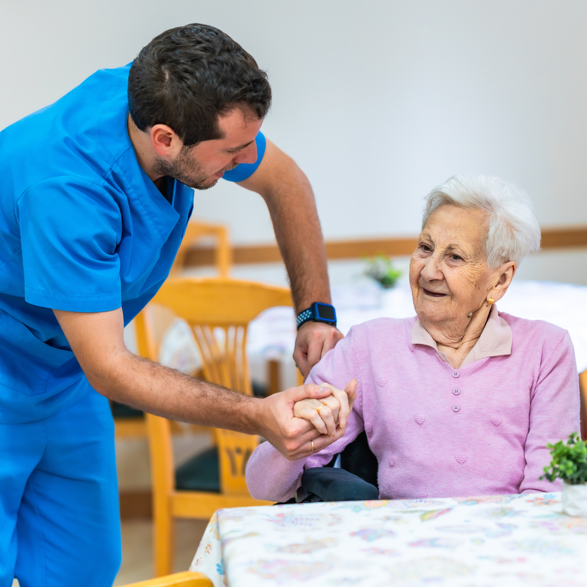 A young male healthcare worker in blue scrubs holding hands and talking with an elderly woman with white hair, seated at a dining table in a care facility.