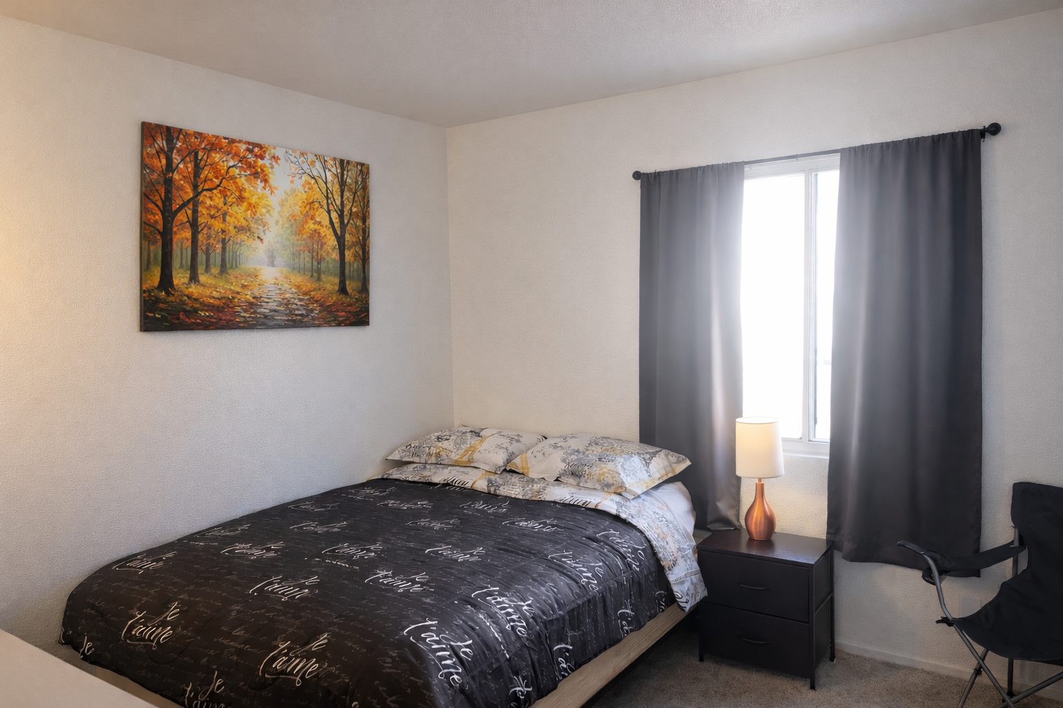 A bedroom with a bed covered in black and white with script words, pillows, a nightstand with a lamp, dark curtains, a window, and a painting of a forest path with orange trees.