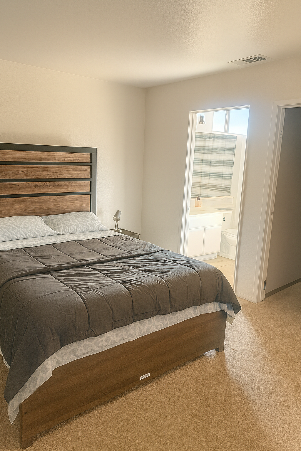A bedroom with a bed, nightstand, and lamp adjacent to a bathroom with a window, sink, and toilet.