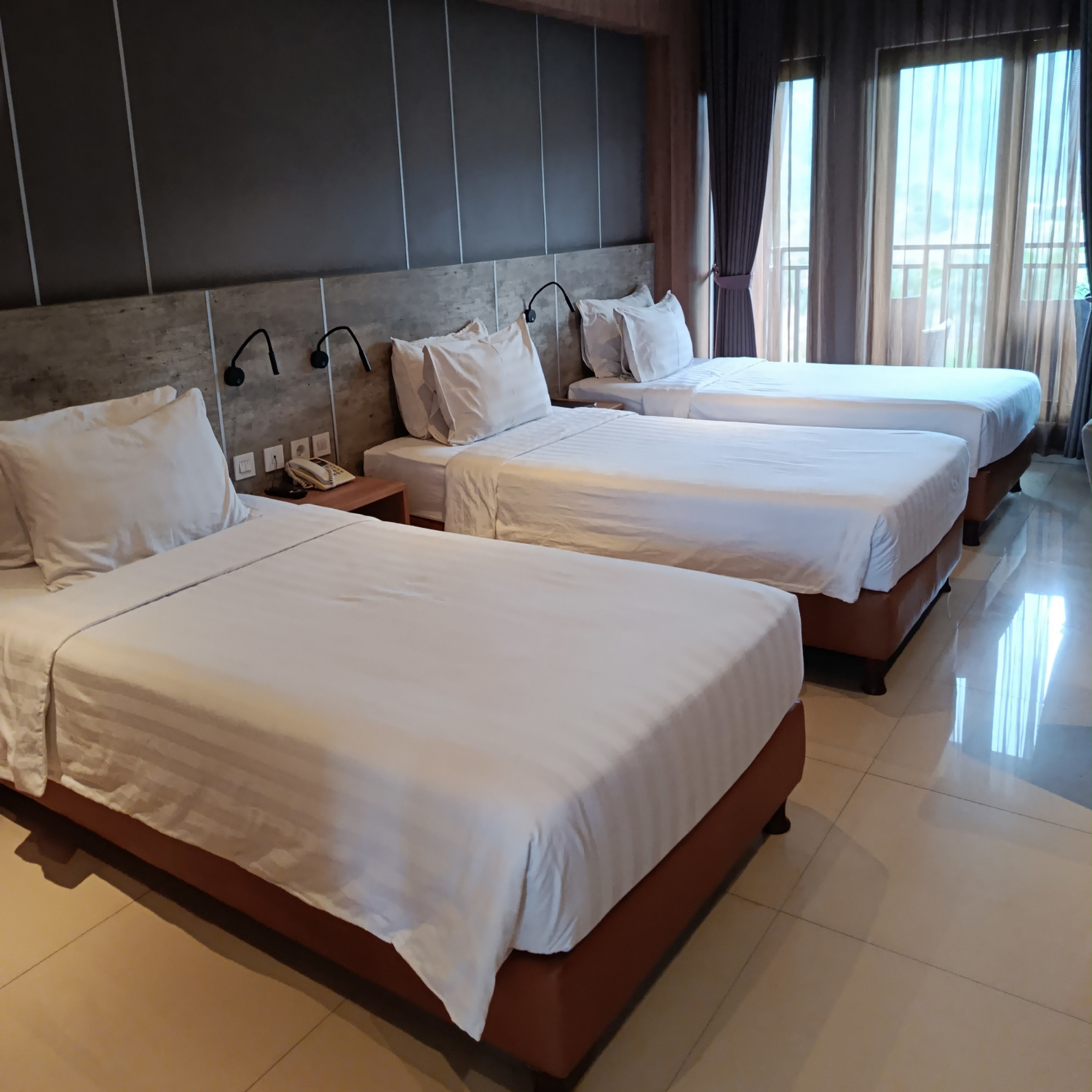 A hotel room with three single beds, each with white linens and pillows. There are reading lights above each bed, and a nightstand with a telephone. Large windows with curtains let in natural light, and the room has shiny tile floors.