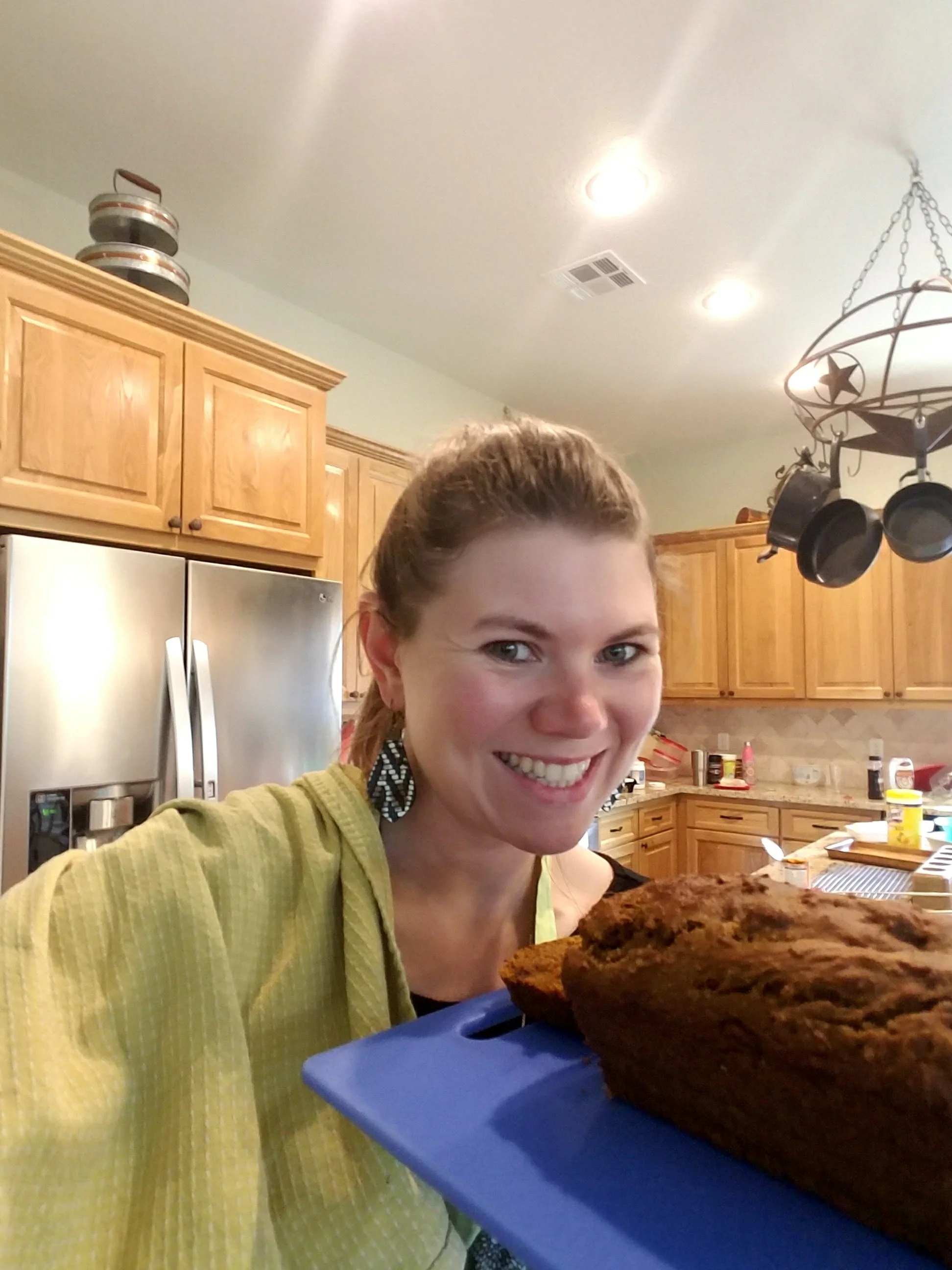 A woman smiling and holding a tray with a large loaf of bread in a kitchen.