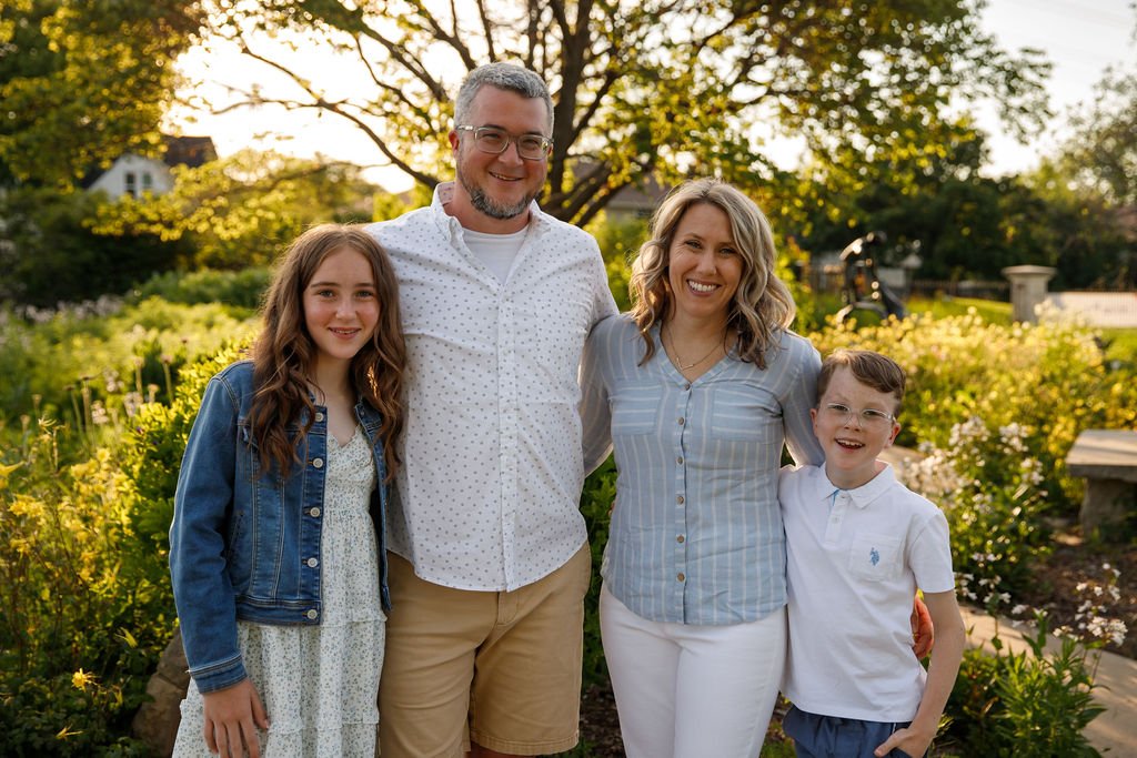 A smiling family of four standing outdoors in a garden during sunset. The family includes a father, mother, and two children, a girl and a boy. They are dressed casually, enjoying a sunny day with trees and flowers in the background.