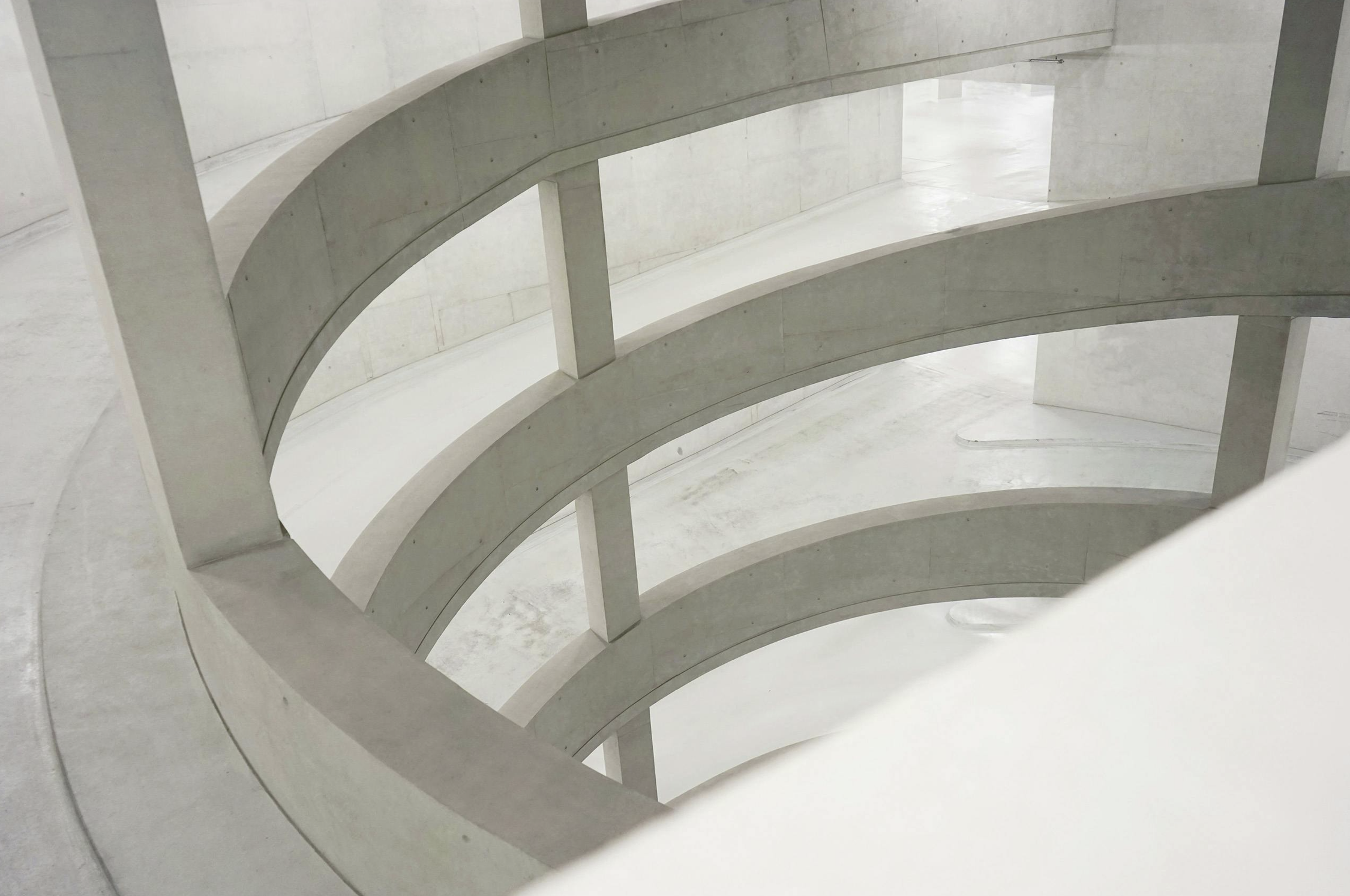 View of a modern, concrete spiral staircase with multiple levels visible from above, in a minimalist architectural setting.