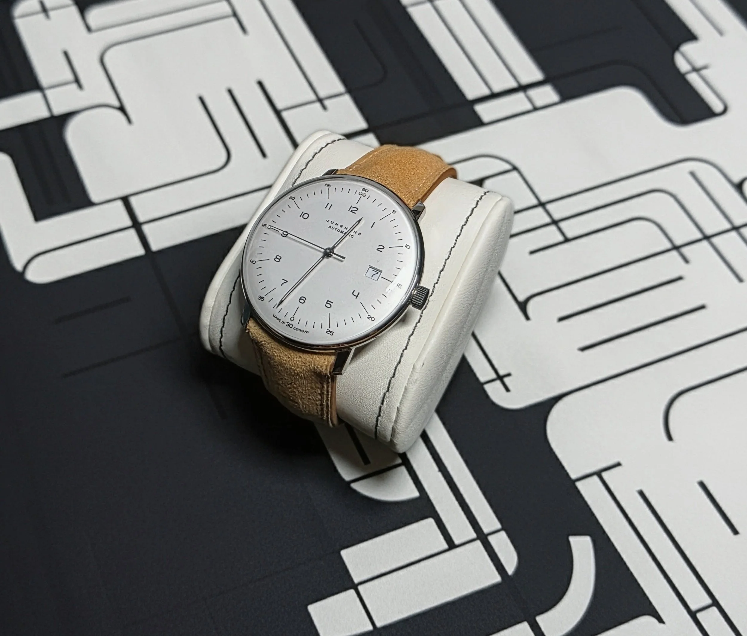 A wristwatch with a white dial and beige leather strap sitting on a white cushion, placed on a black surface with white abstract line designs.