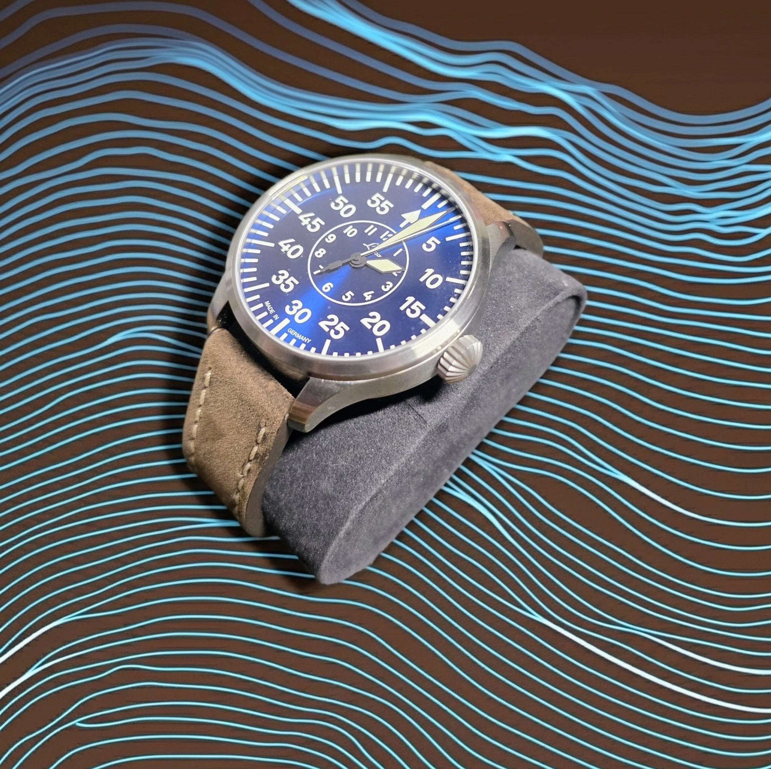 A wristwatch with a blue dial and beige strap resting on a textured gray cushion, against a background with blue wavy lines on a brown surface.