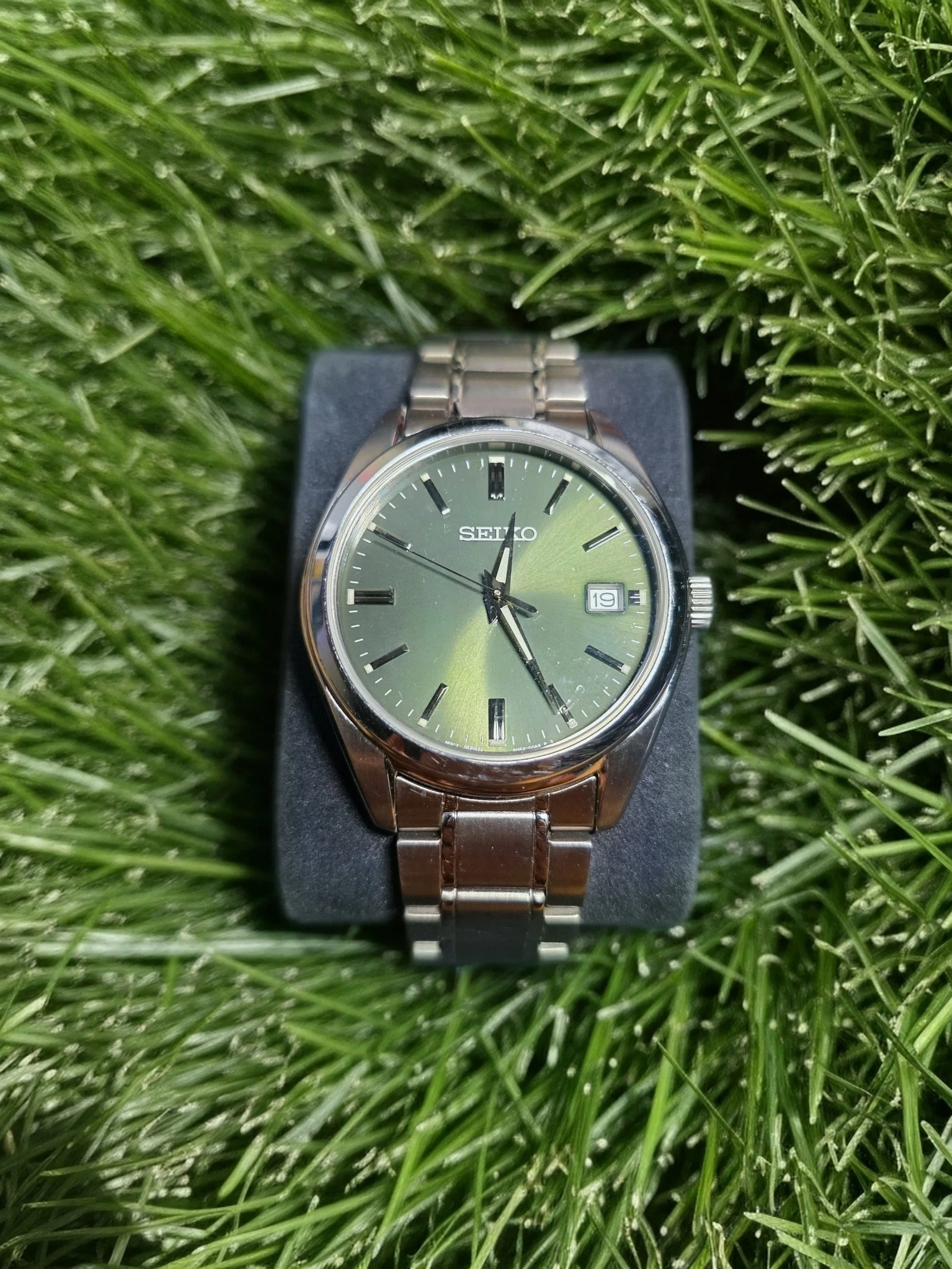 A silver Seiko wristwatch with a green face and silver band, placed on a black cushion amidst green grass.