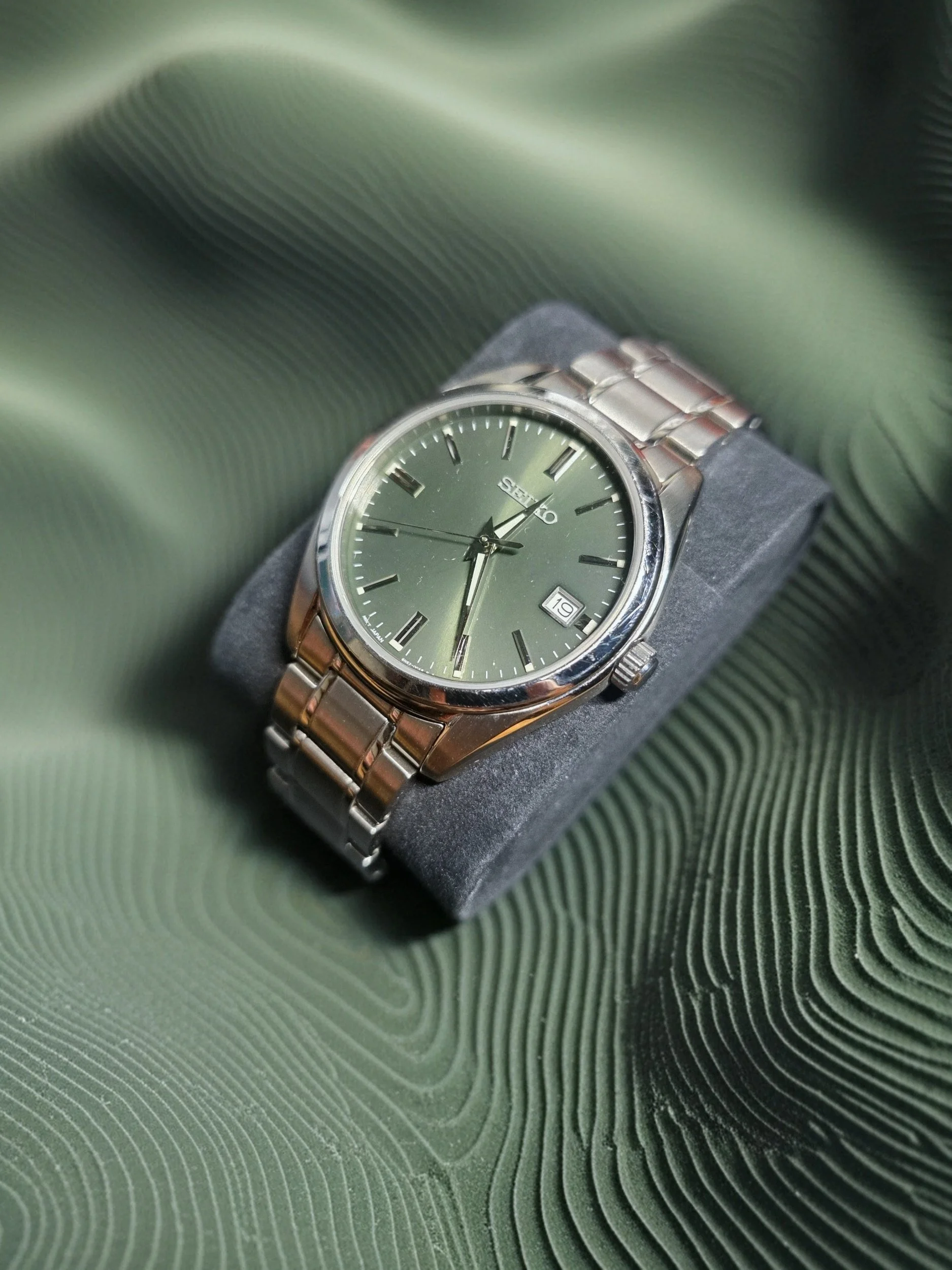 A silver Seiko wristwatch with a green face, silver hour markers, and a date window at 3 o'clock, resting on a dark cushion with a textured green background.