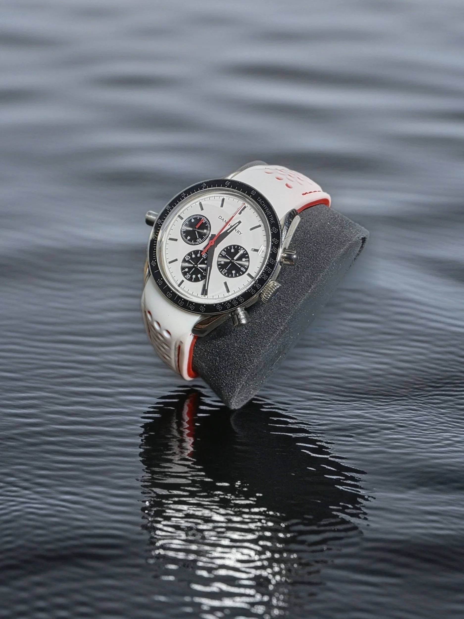 A wristwatch with a white strap, black bezel, and white dial showing subdials, placed on a foam cushion on a reflective water surface.