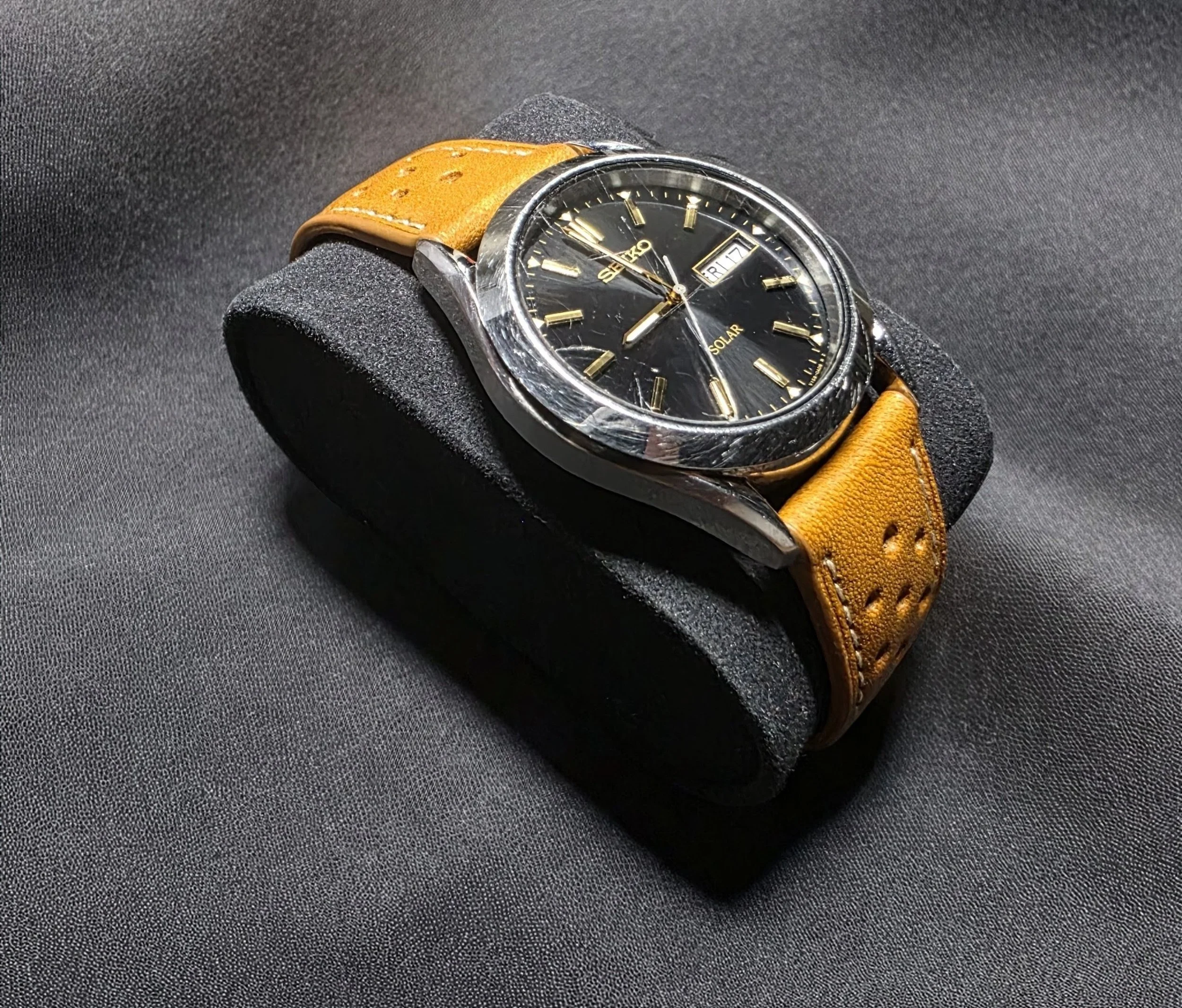 A vintage Seiko solar wristwatch with a black dial, gold markers, and a day/date display, placed on a black cushioned stand with a tan leather strap, against a textured dark background.