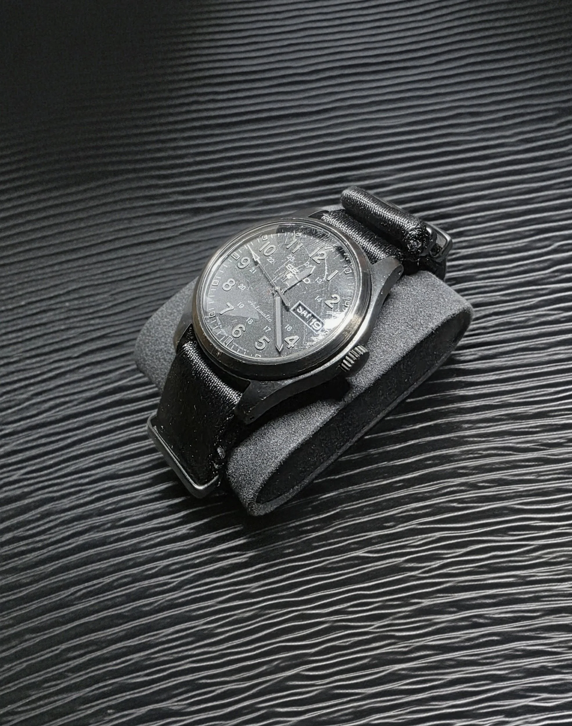 A black analog wristwatch with a black leather strap, placed on a foam cushion on a textured black surface.