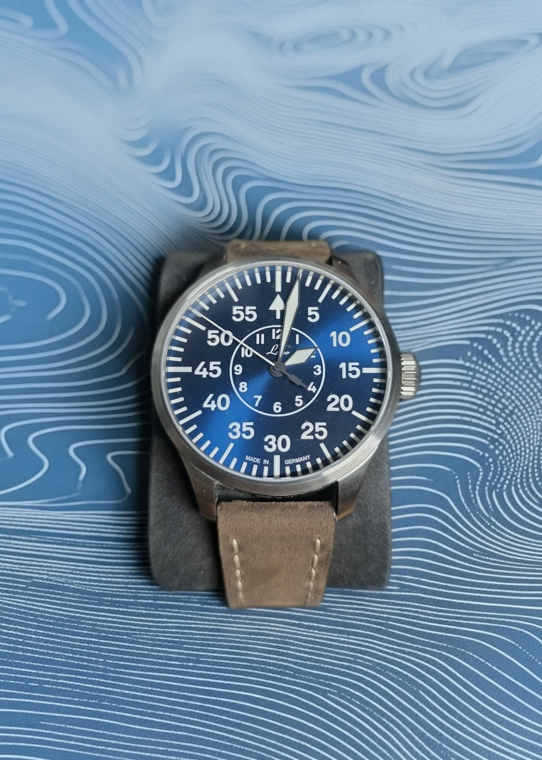A wristwatch with a blue dial, silver case, and beige leather strap resting on a blue patterned surface.
