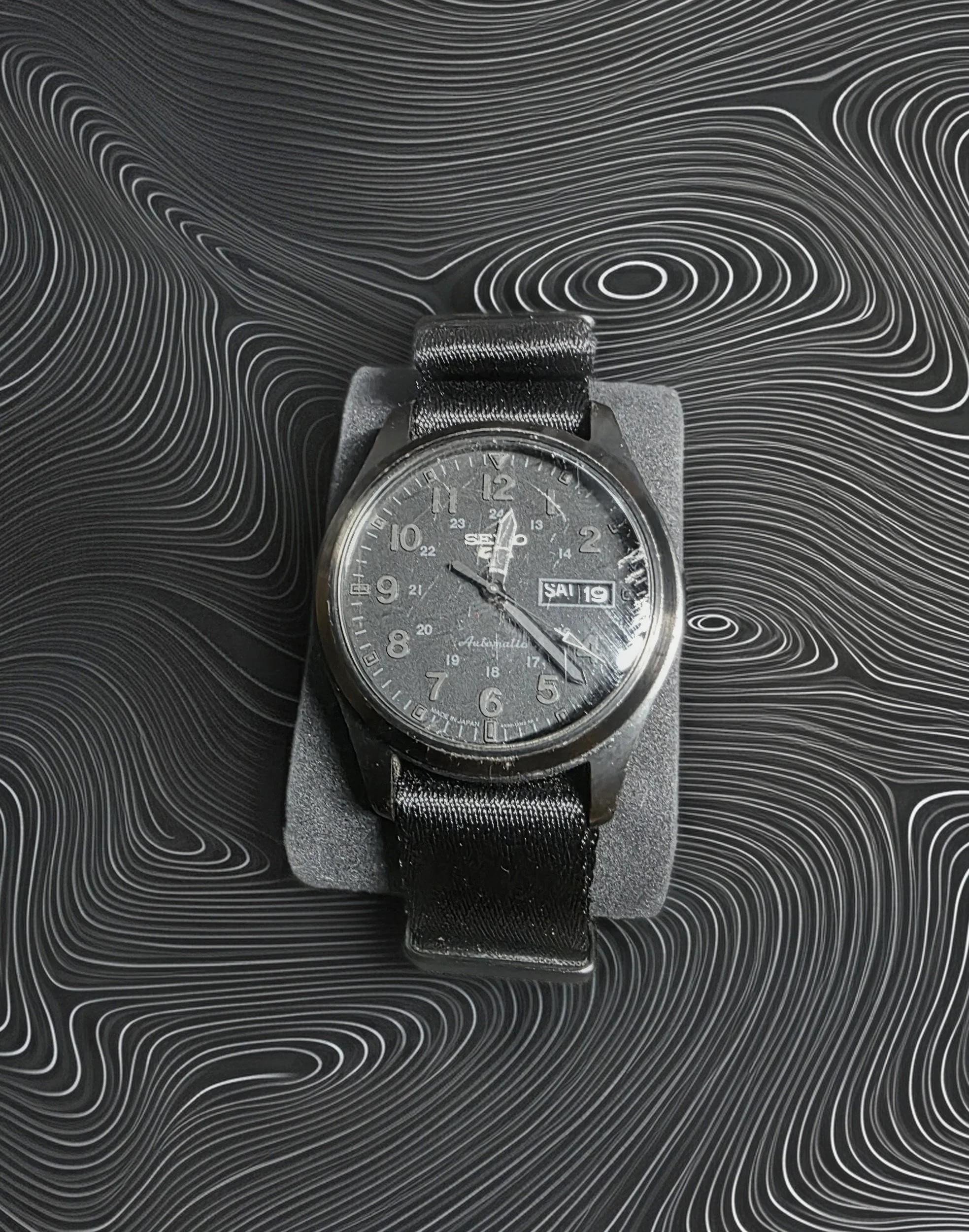 Black generic analog wristwatch with day and date display, placed on a foam cushion on a black textured surface.