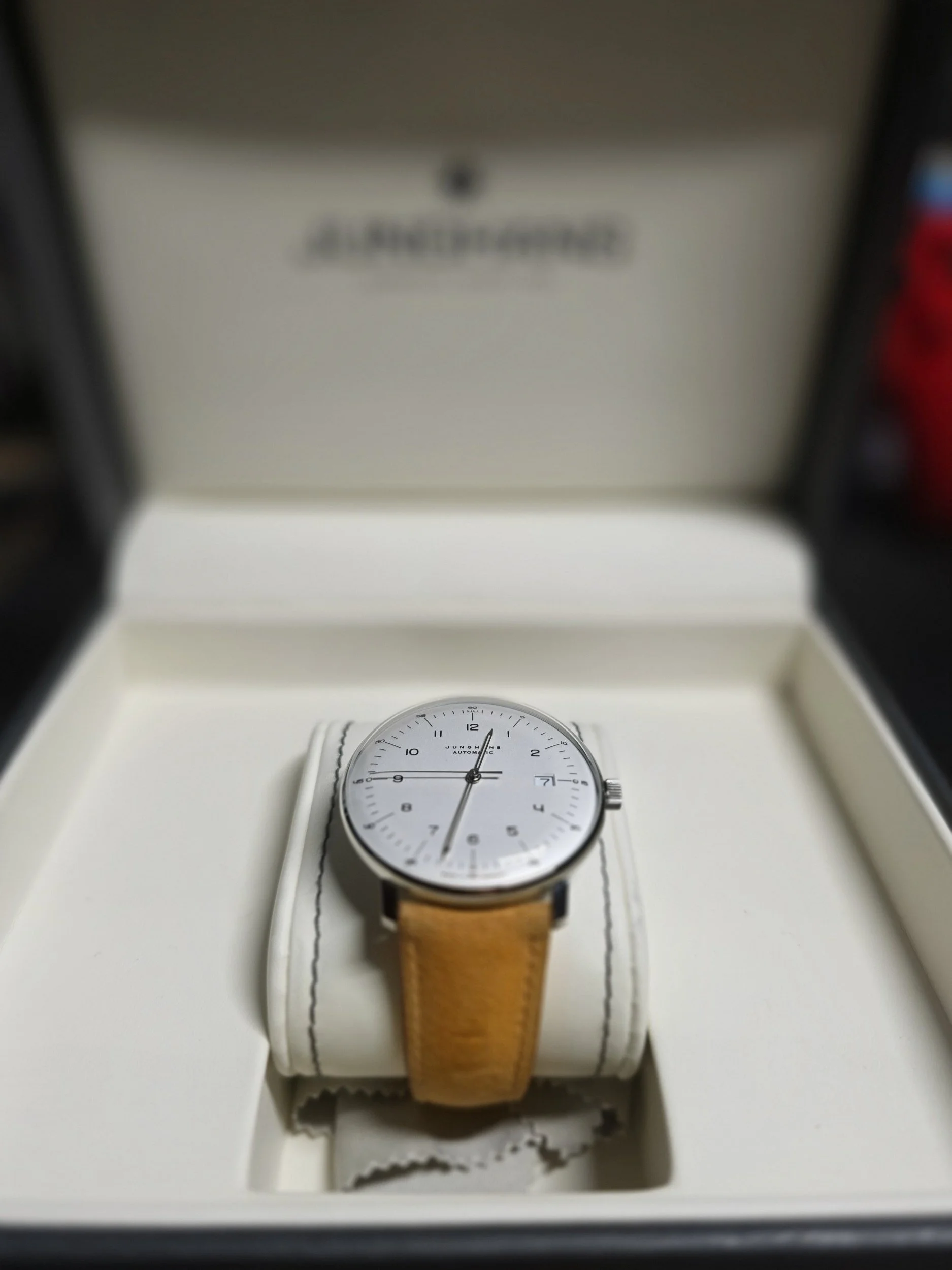 A white wristwatch with a brown leather strap displayed inside a white box, with the watch face showing the time and a date window at 3 o'clock.