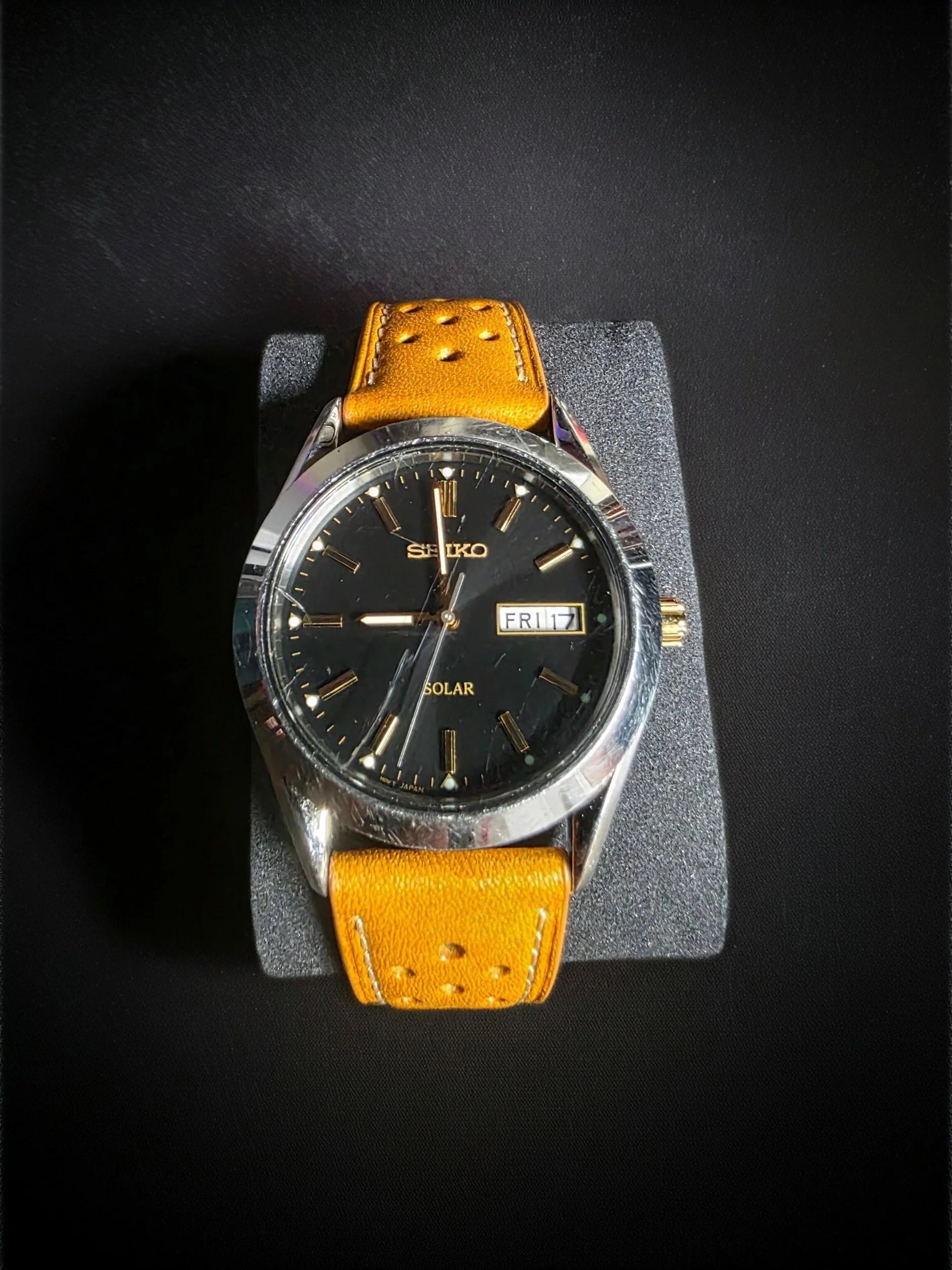 Seiko solar wristwatch with black dial, gold hour markers, and an orange leather strap, displayed on a black foam cushion.
