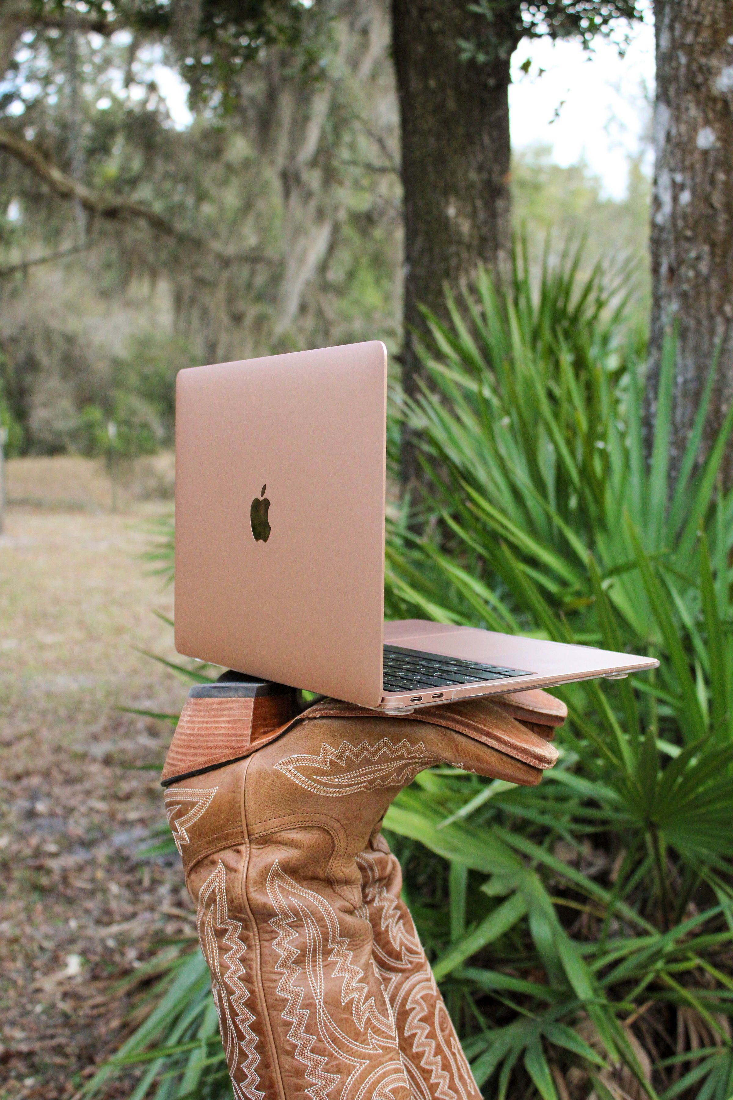 A pink Apple MacBook placed on a brown leather glove, held upright in an outdoor wooded area with green foliage and trees in the background.