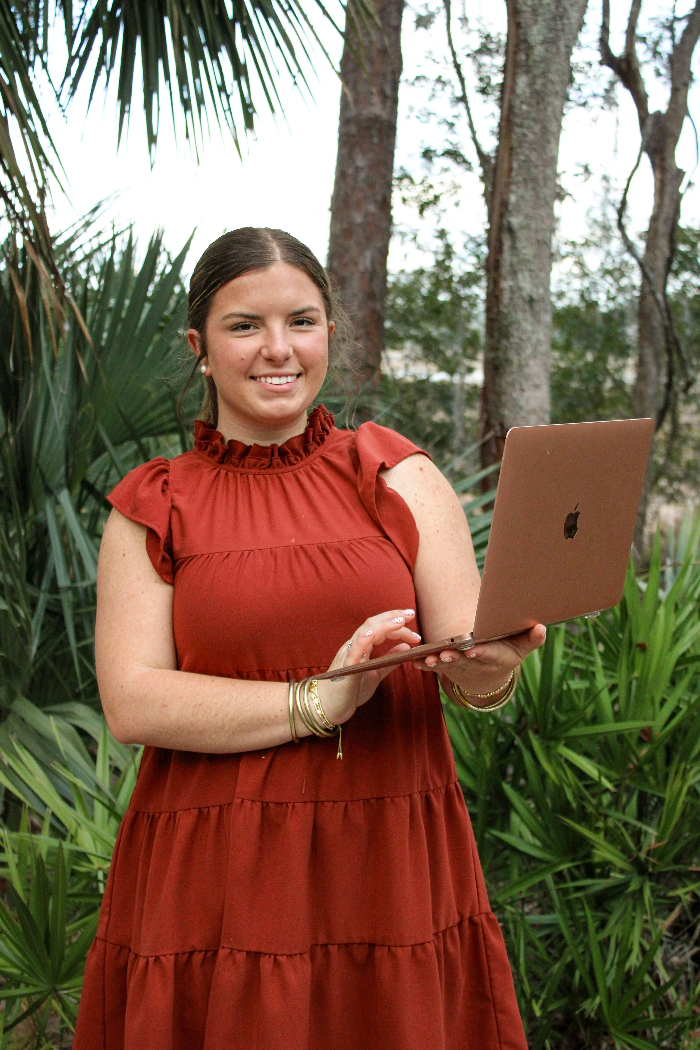A young woman with dark brown hair, wearing a red ruffled dress and gold jewelry, is standing outdoors in a wooded area with trees and green foliage, holding a rose gold MacBook laptop and smiling at the camera.