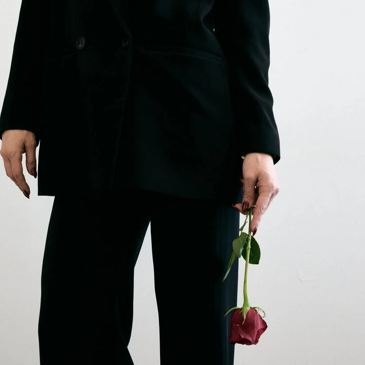 Person dressed in black holding a red rose with green leaves, standing against a plain white background.
