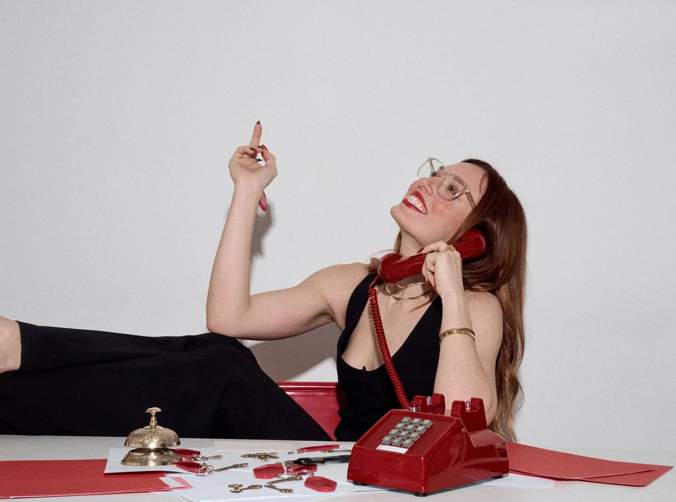 A woman with long red hair, wearing glasses and red lipstick, is talking on a red rotary telephone. She is smiling, raising her right hand with her index finger pointing up, and is wearing a black sleeveless top. There is a white desk with various jewelry items, a decorative clock, and a red folder in front of her.