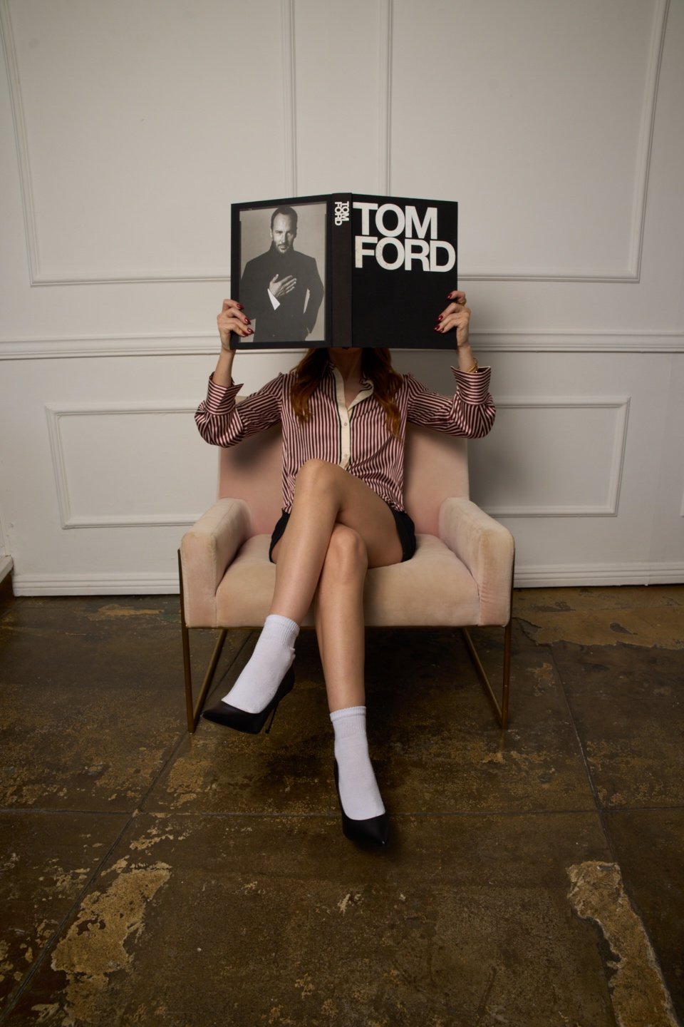 Person sitting on a pink chair, wearing black shorts, white socks, and black shoes, holding a large black and white Tom Ford magazine in front of their face. The magazine features a black and white photograph of Tom Ford.