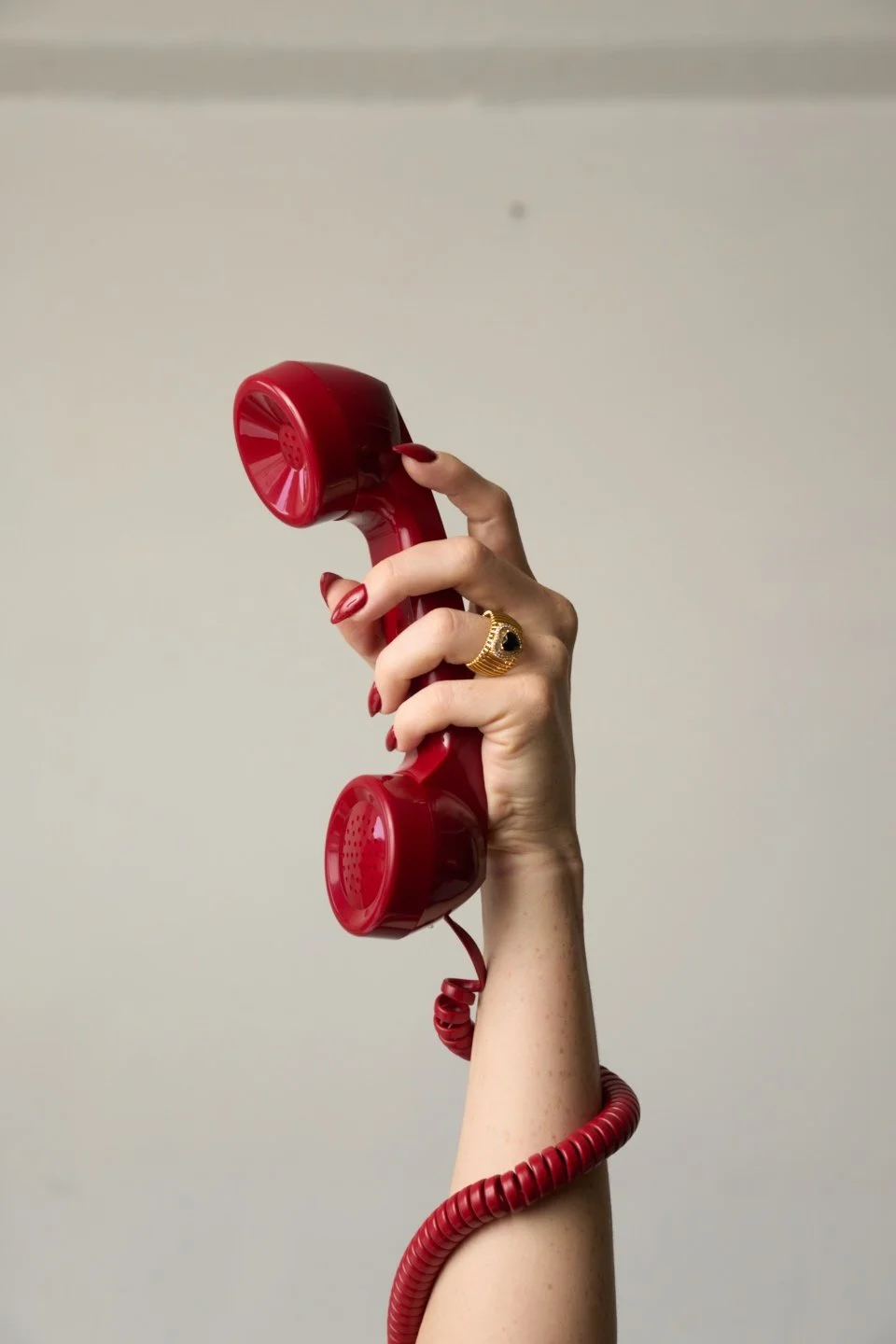 A person with red manicured nails holding a classic red rotary phone receiver with a gold ring finger, against a plain light background.