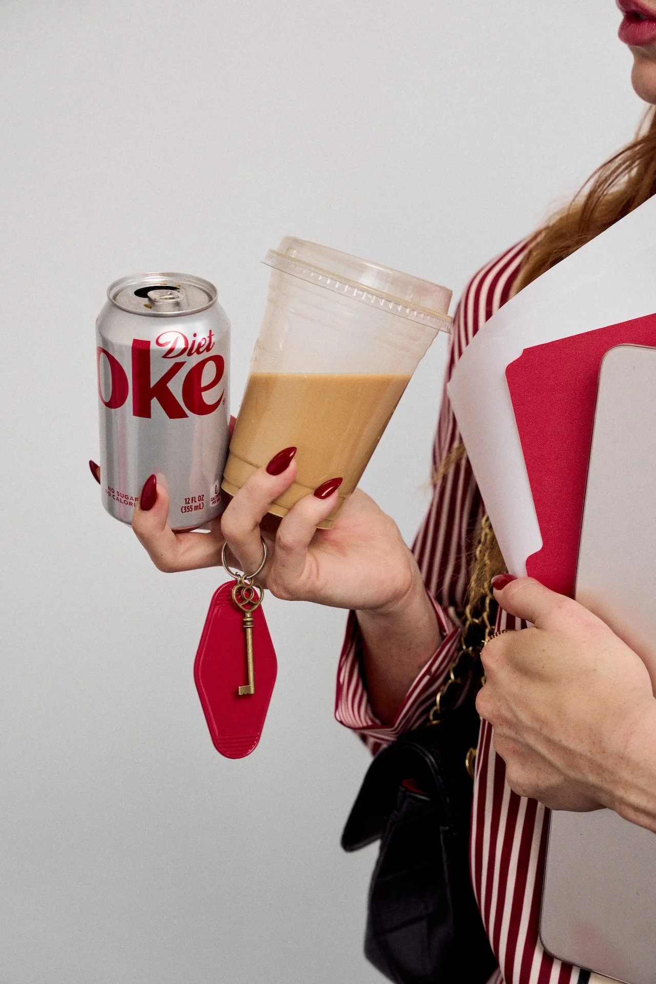 Person holding a Diet Coke can, a frappuccino, and a folder, with red lipstick and red nails, against a plain white background.
