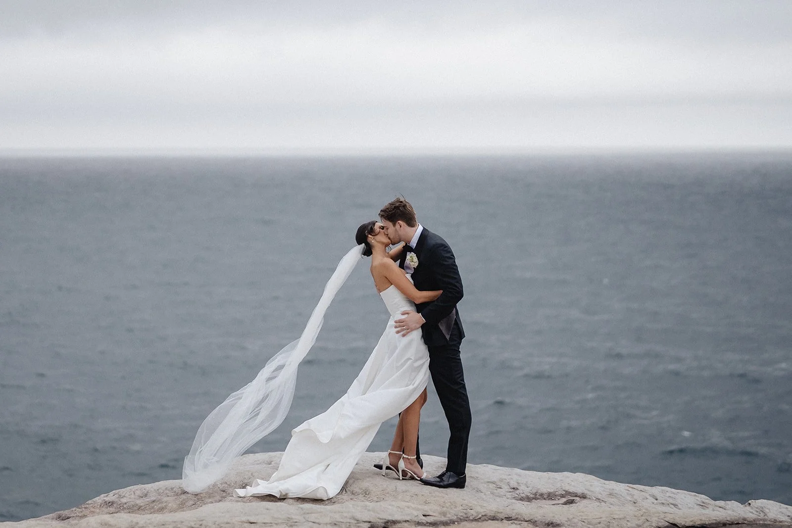 A bride and groom kissing on a rocky outcrop by the ocean, with the bride in a white wedding gown and veil, and the groom in a black suit and tie.
