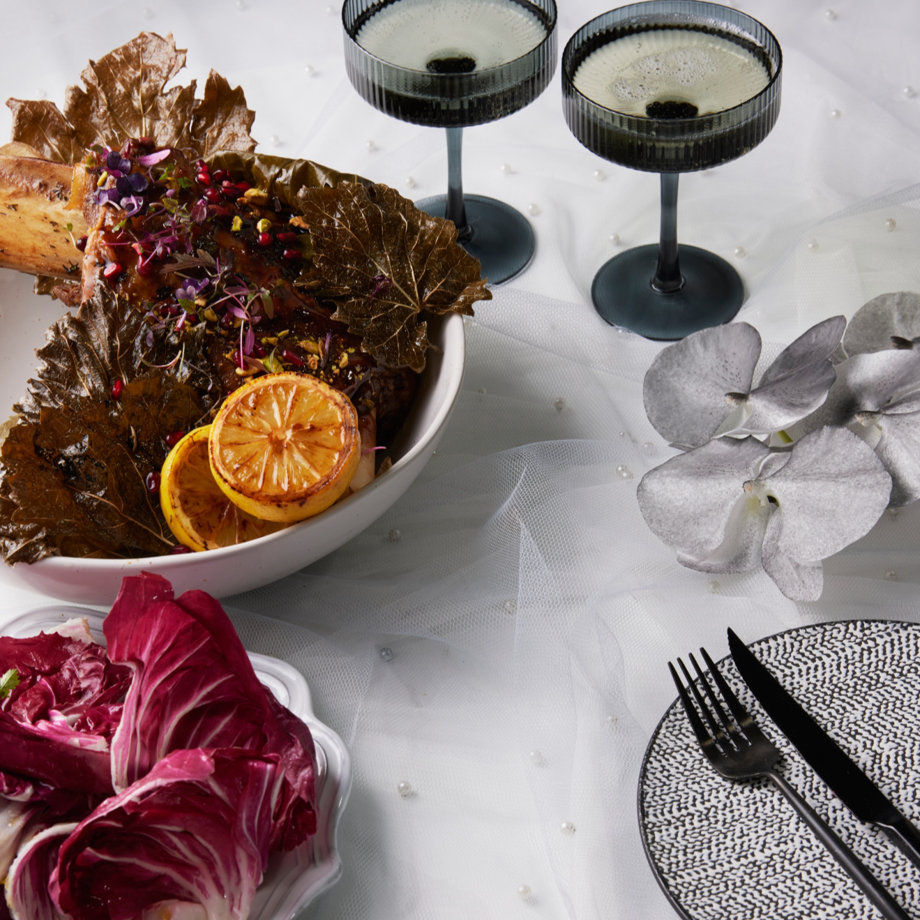 Decorative table setting with a bowl of roasted vegetable salad, two black wine glasses filled with white wine, a side dish of radicchio, a white flower, and a patterned plate with cutlery, all on a white tablecloth.